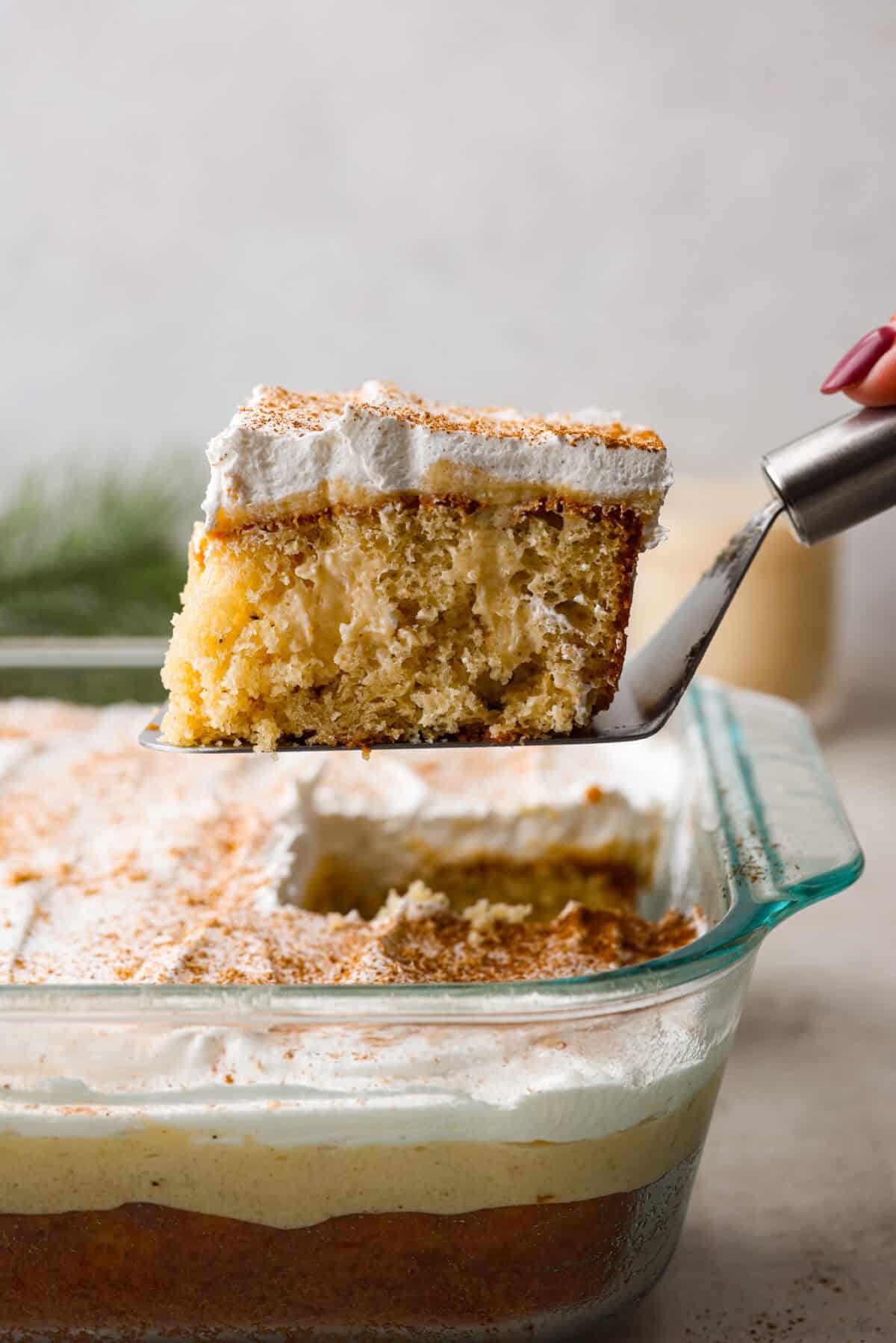 Eggnog Poke Cake could not be more perfect for the holiday season! It combines the rich flavor of eggnog with the soft, moist texture of a classic poke cake. Add fluffy whipped topping and a sprinkle of nutmeg and cinnamon to really wow your family! Side shot of a piece of eggnog poke cake being lifted out of the baking dish.