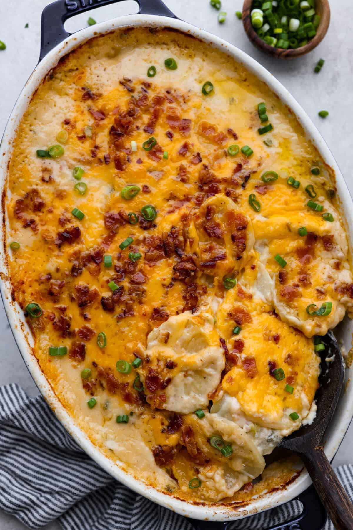 Loaded scalloped potatoes are rich, creamy, and "loaded" with flavor! This recipe combines tender potatoes baked to perfection in a velvety sauce topped with crispy bacon, green onions, and melty cheese. Overhead shot of loaded scalloped potatoes in a baking dish.