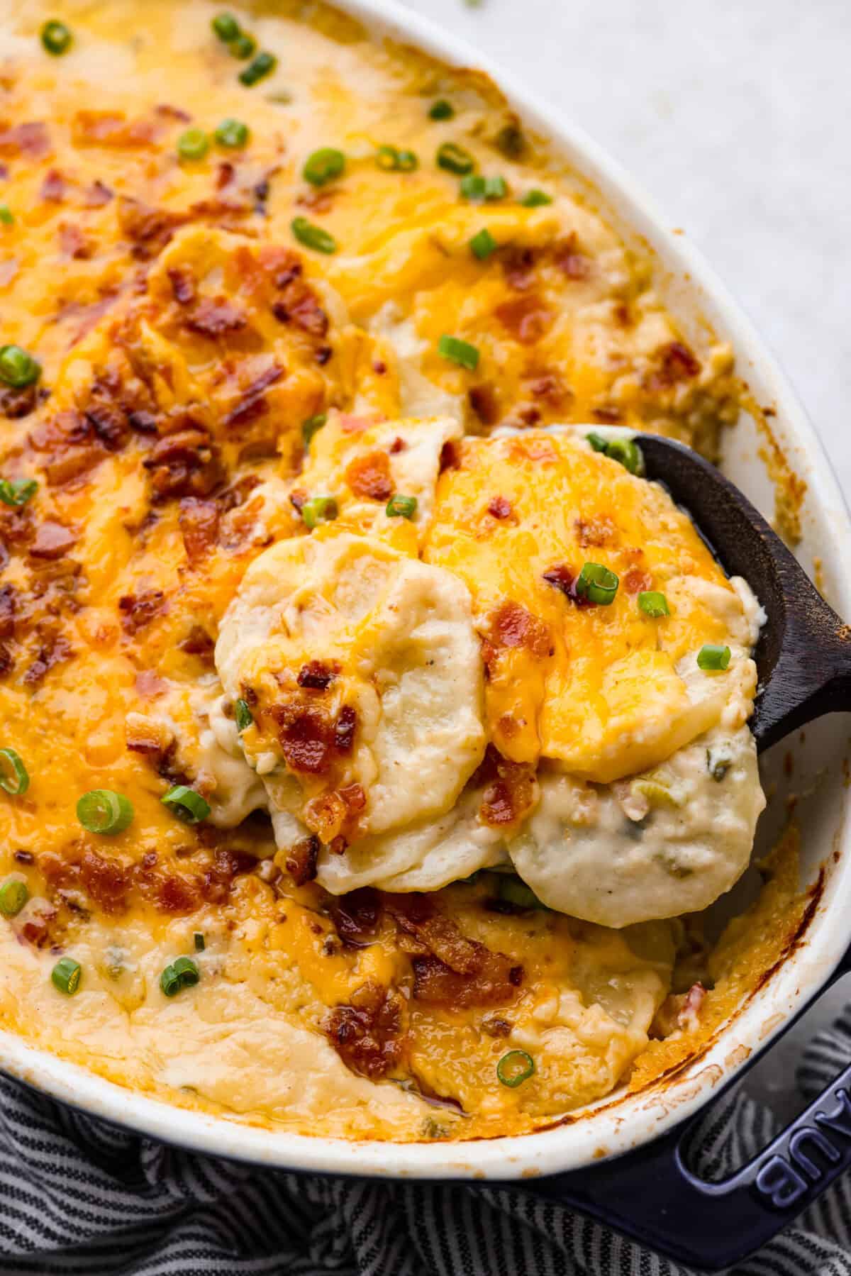 Loaded scalloped potatoes are rich, creamy, and "loaded" with flavor! This recipe combines tender potatoes baked to perfection in a velvety sauce topped with crispy bacon, green onions, and melty cheese. Angle shot of someone scooping out a serving of loaded scalloped potatoes.
