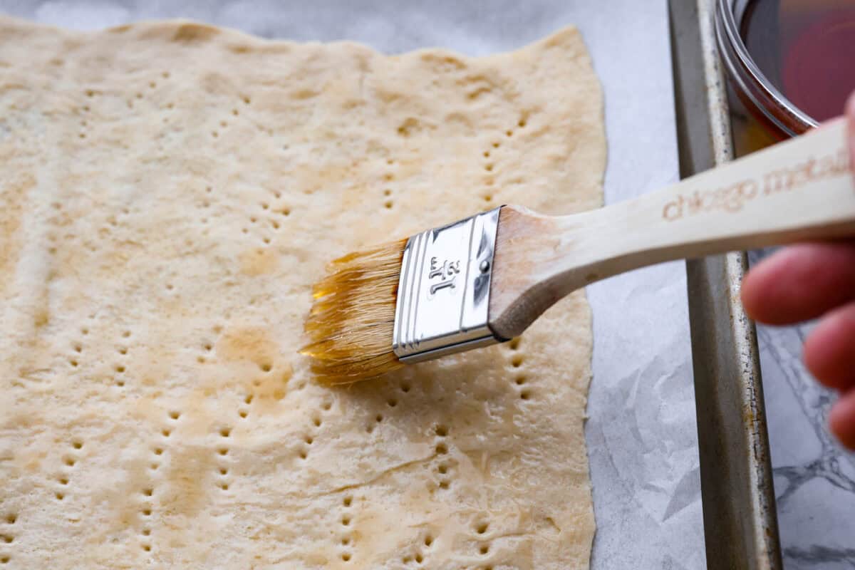 Maple Bacon Crack is as delicious and addicting as it sounds! It has flaky pastry dough drizzled in a sweet maple sauce and is loaded with crispy bacon pieces! It's the perfect sweet and salty snack! Angle shot of someone spreading maple syrup on the crescent dough sheet.