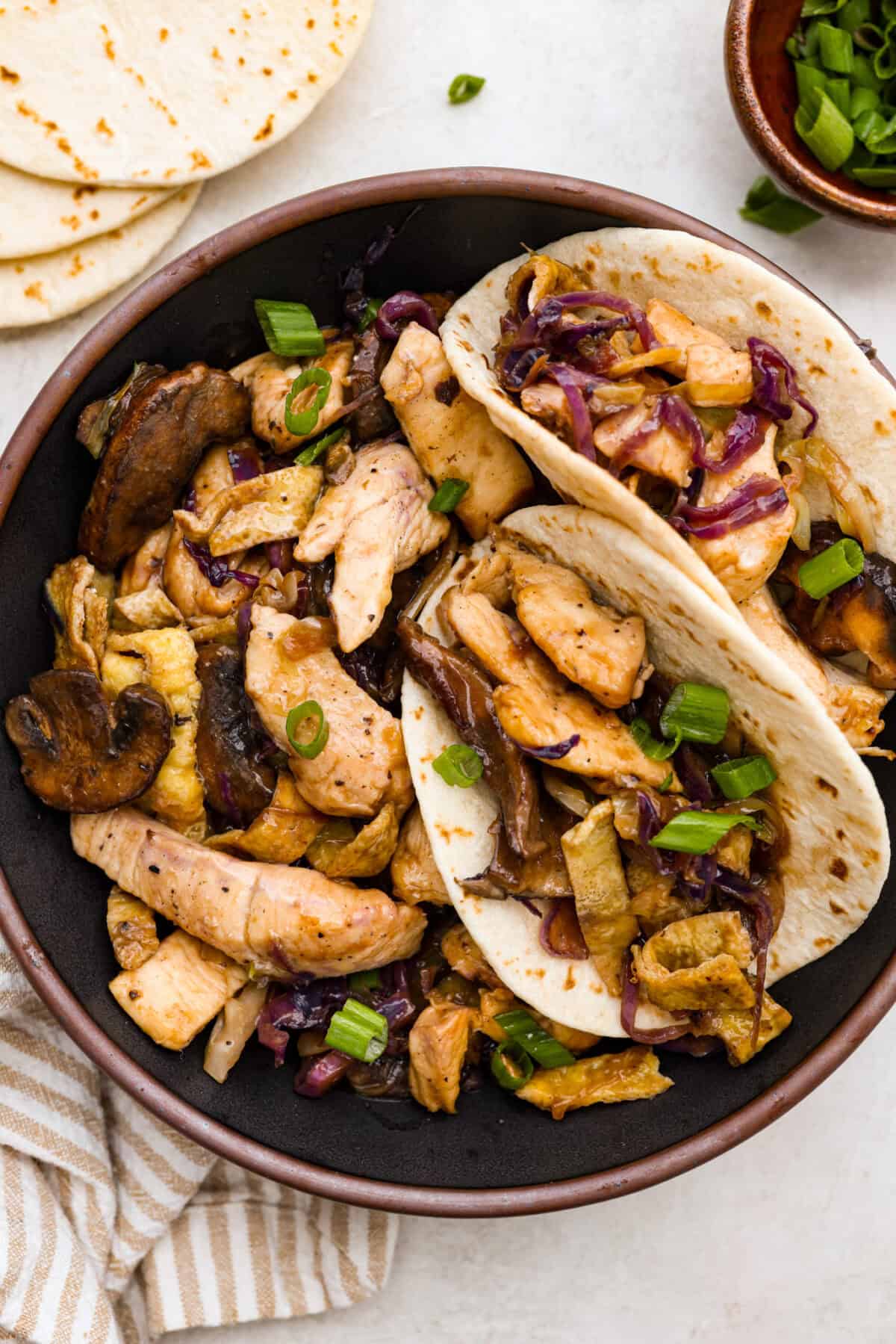 Moo Shu Chicken is a takeout favorite that’s so easy to make at home! This flavorful dish combines tender strips of chicken, mushrooms, and cabbage with a sweet and savory sauce. It’s quick, packed with vibrant flavors, and the perfect weeknight meal! Overhead shot of plated Moo Shu chicken with some in tortillas.