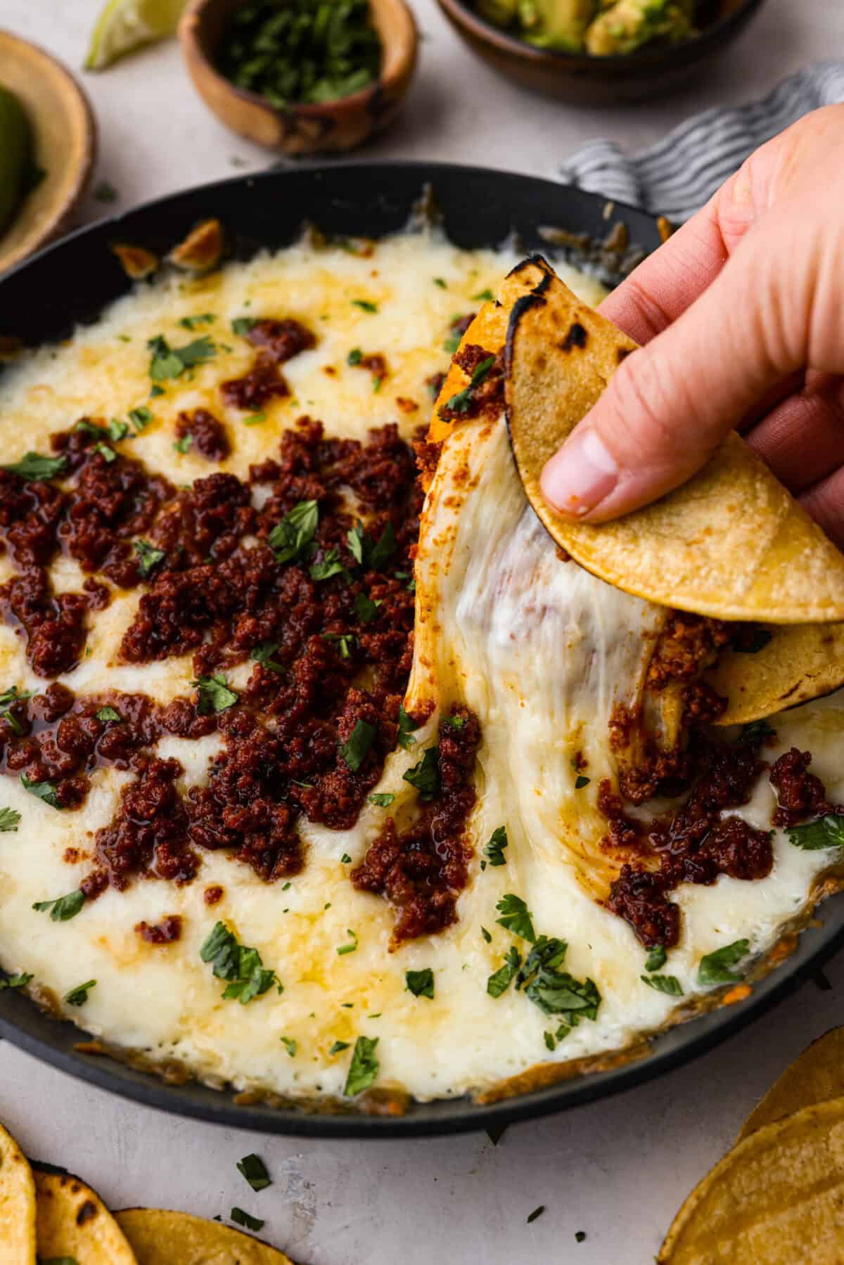 Queso Fundido is a delicious cheesy appetizer with a kick from the Mexican chorizo. It's loaded with buttery, melty cheeses and covered in spicy chorizo sausage. Close up shot of someone pulling up some queso fundido in a corn tortilla.