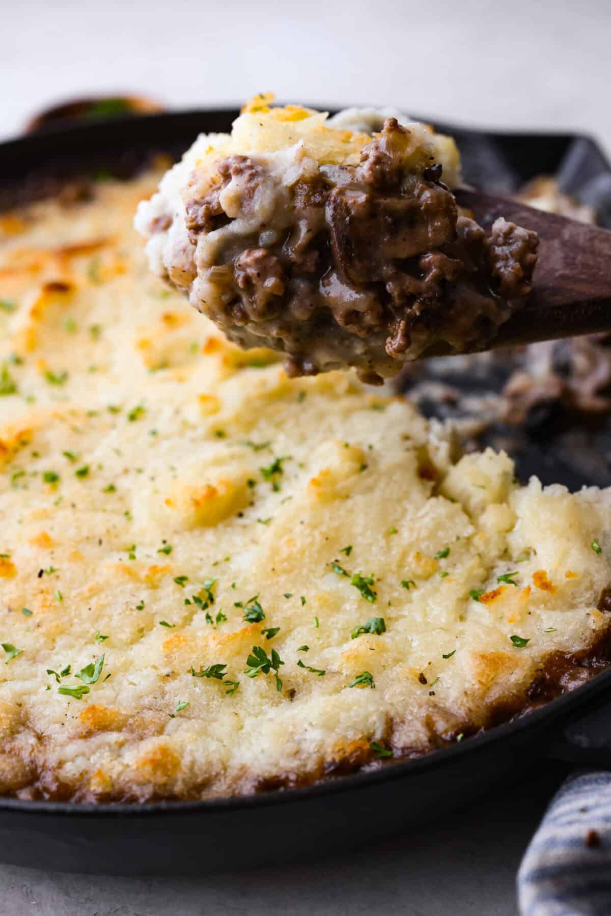 Salisbury Steak casserole is a warm and comforting savory dish. It has a thick beef gravy, tender mushrooms, and seasoned hamburger topped with rich, creamy potatoes. Side shot of someone lifting a scoop of the salisbury steak casserole out of the skillet.
