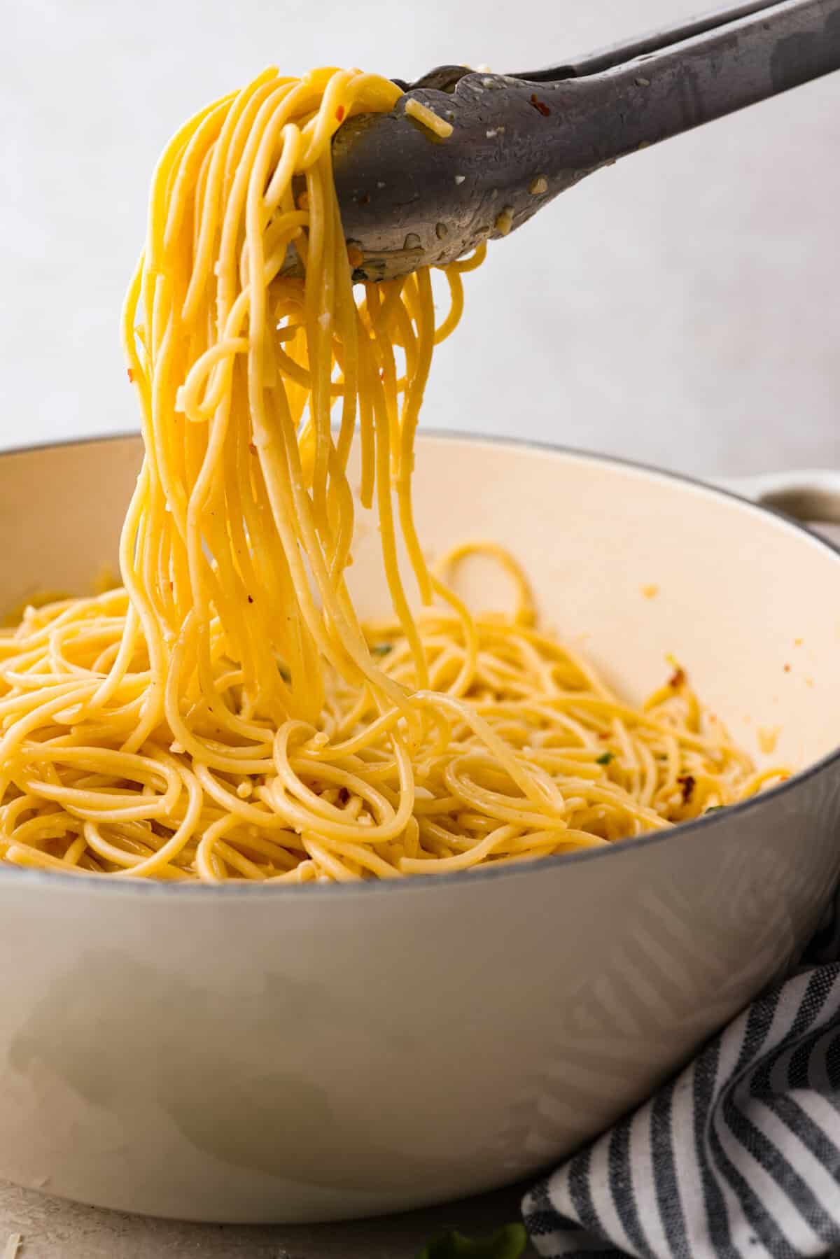Spaghetti Aglio e Olio uses simple ingredients to create a dish bursting with flavor! With just garlic, olive oil, chili flakes, pasta, and parmesan cheese, this classic Italian recipe is a go-to for a tasty meal in less than 30 minutes. Side shot of someone lifting some of the spaghetti aglio e olio from the pot with tongs.