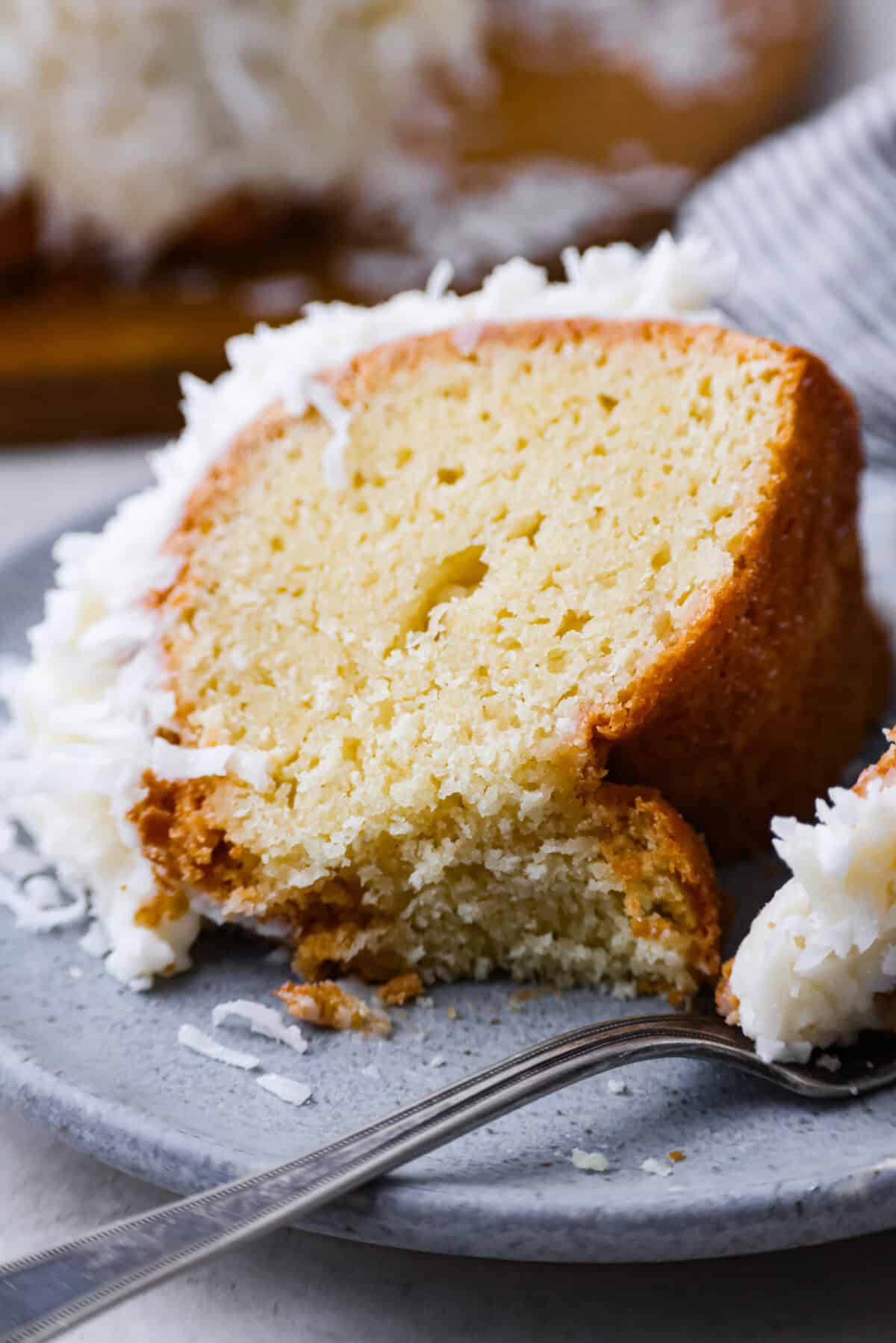 Tom Cruise coconut cake is a dense pound cake with coconut flavor and silky white chocolate throughout. The indulgent frosting is a coconut cream cheese buttercream with sweetened coconut on the outside. It's a coconut lover's dream—but a super sweet one. Close up shot of a piece of plated Tom Cruise coconut cake.