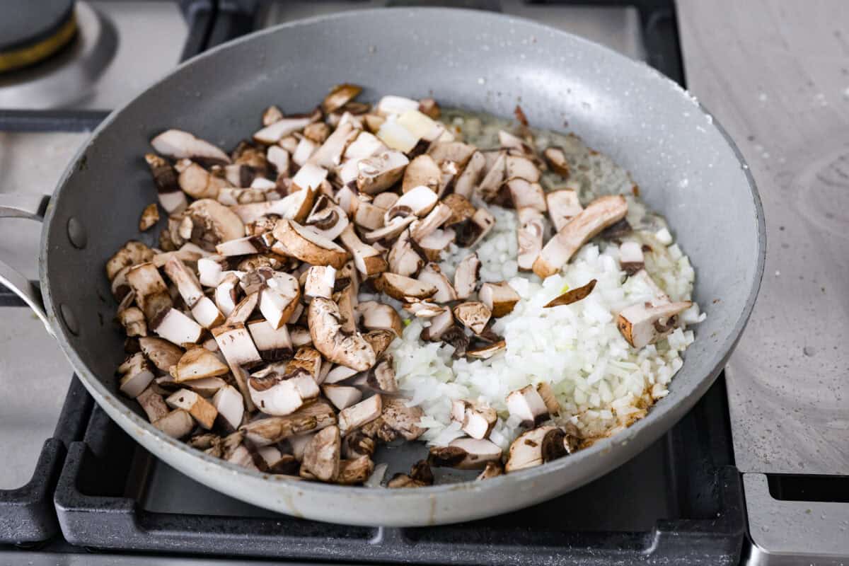 This Chicken Gloria is moist, perfectly baked chicken with a slightly seared crust. It cooks in a creamy sauce that is slightly tangy and full of flavor. It's topped with gooey cheese and makes a very satisfying meal! Angle shot of onions garlic and mushrooms cooking in butter in a skillet.