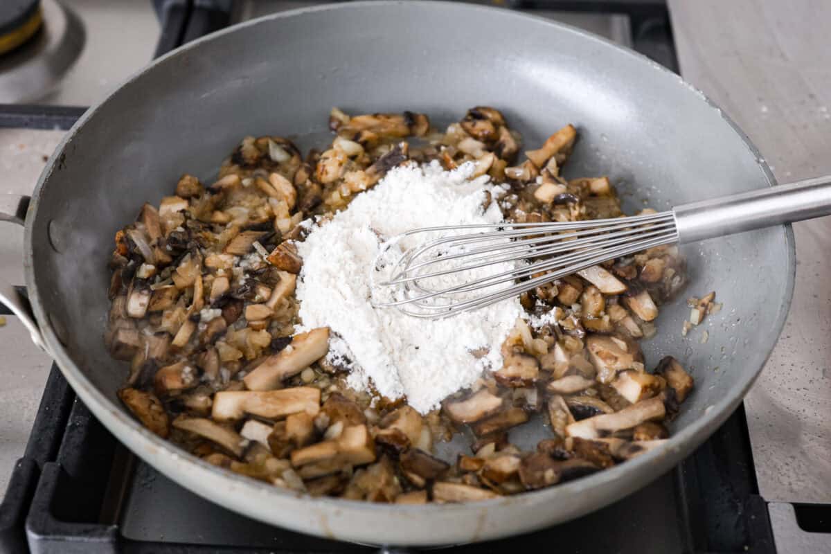 This Chicken Gloria is moist, perfectly baked chicken with a slightly seared crust. It cooks in a creamy sauce that is slightly tangy and full of flavor. It's topped with gooey cheese and makes a very satisfying meal! Angle shot of someone whisking the flour into the mushrooms and onions.