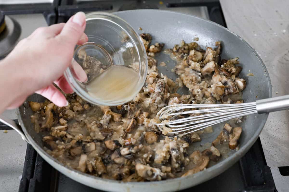 This Chicken Gloria is moist, perfectly baked chicken with a slightly seared crust. It cooks in a creamy sauce that is slightly tangy and full of flavor. It's topped with gooey cheese and makes a very satisfying meal! Angle shot of someone pouring chicken broth into the skillet with the flour and mushroom mixture.