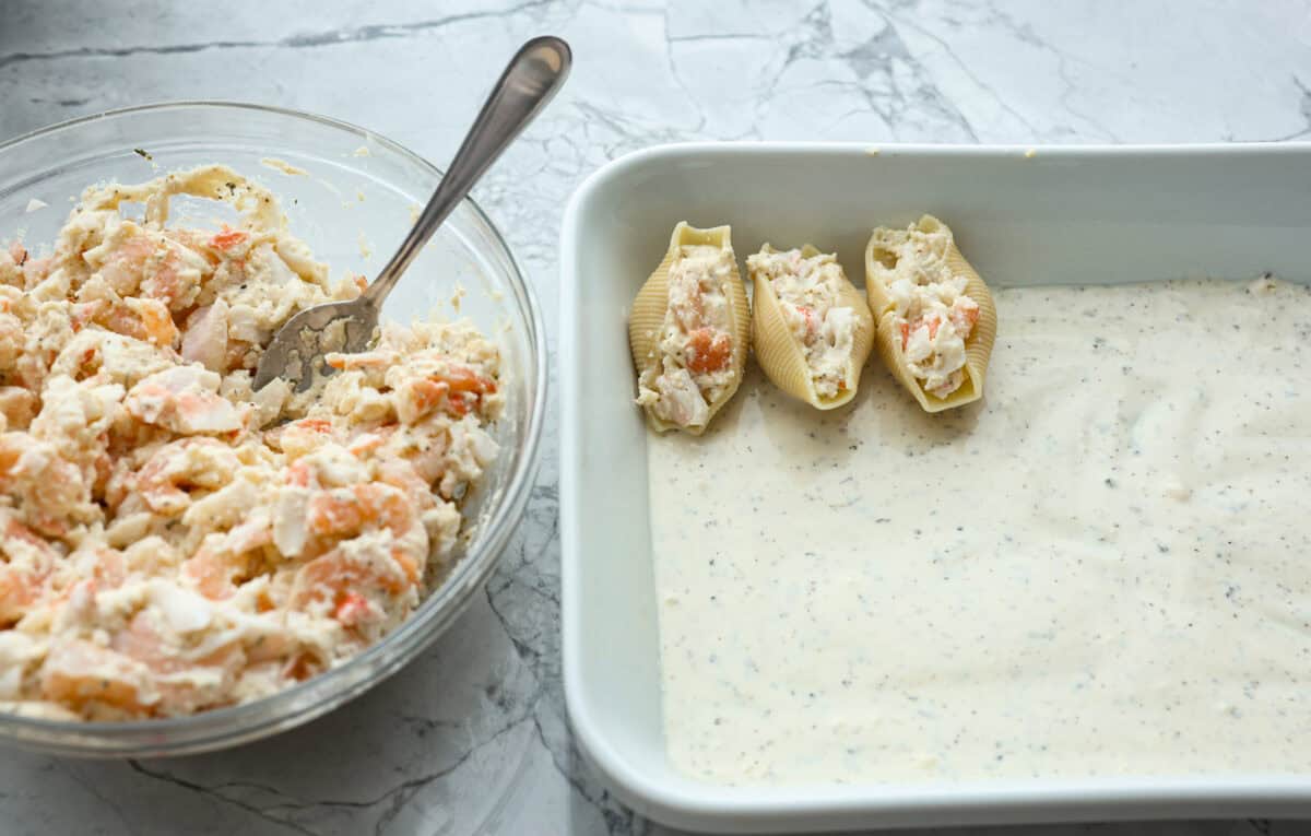 Seafood Stuffed Shells is a comforting meal that's sure to impress! Jumbo pasta shells are generously filled with a creamy, savory seafood filling and coated in a rich Alfredo-based sauce. Finish with a crispy crumb topping for an irresistible dinner! Angle shot of Alfredo sauce in the bottom of a baking dish with a few seafood stuffed shells on top of the sauce in the pan.