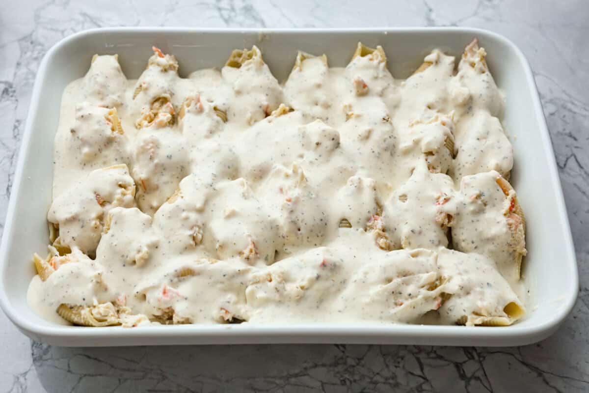 Seafood Stuffed Shells is a comforting meal that's sure to impress! Jumbo pasta shells are generously filled with a creamy, savory seafood filling and coated in a rich Alfredo-based sauce. Finish with a crispy crumb topping for an irresistible dinner! Overhead shot of all the pasta shells stuff and covered in Alfredo sauce.
