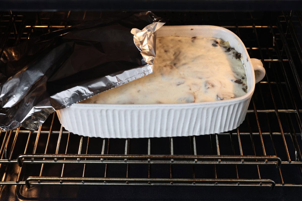 This creamy, cheesy mushroom lasagna is a delicious twist on the classic. Instead of traditional meat and red sauce, this recipe features layers of garlicky mushrooms and a rich bechamel sauce that brings incredible depth to every bite. Side shot of mushroom lasagna cooking in the oven with someone taking the tin foil off.