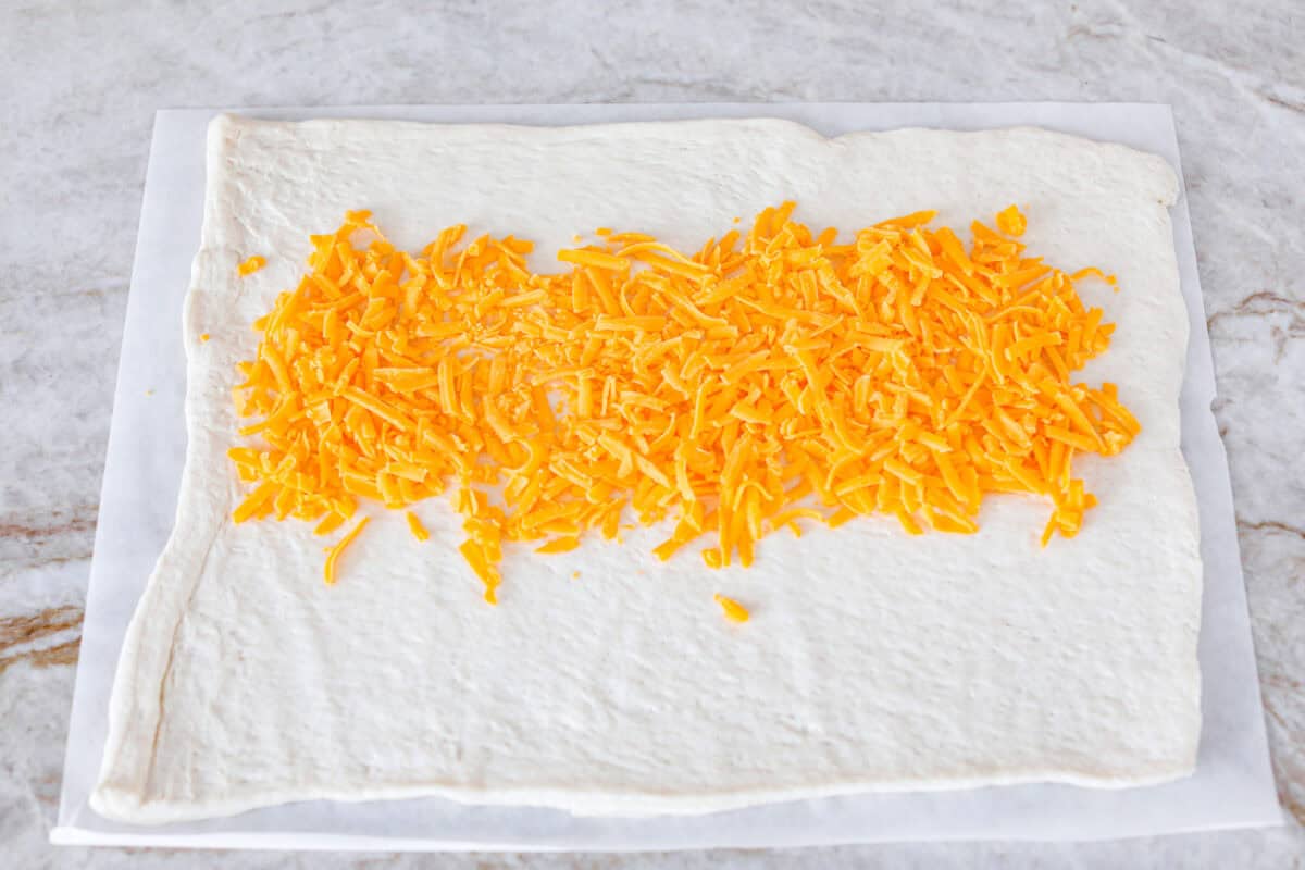 This Breakfast Stromboli is the best morning treat! It's packed with fluffy eggs, savory sausage, crispy bacon, and melty cheese, all wrapped in golden pizza dough. Perfect for holiday brunches or everyday family breakfasts! Overhead shot of the pizza crust dough rolled out onto parchment paper with cheese sprinkled through the middle.