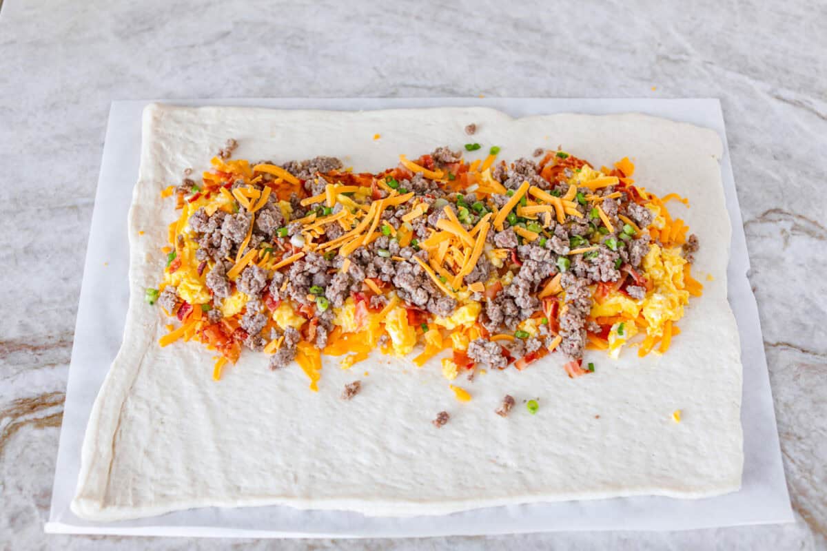 This Breakfast Stromboli is the best morning treat! It's packed with fluffy eggs, savory sausage, crispy bacon, and melty cheese, all wrapped in golden pizza dough. Perfect for holiday brunches or everyday family breakfasts! Angle shot of sausage, bacon, and green onion sprinkled over the cheese.