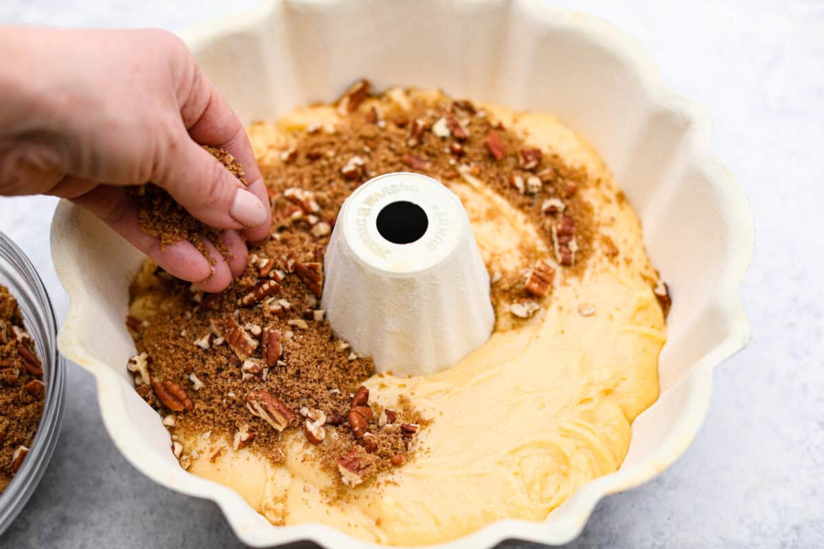 This Sock It to Me Cake is a moist, buttery Southern bundt cake with a delicious cinnamon-sugar pecan layer swirled through the middle. It became popular because of Duncan Hines in the 1960s and 1970s, and you'll understand why once you try it! Overhead shot of someone sprinkling the pecan mixture over the sock it to me cake batter in the bottom of the bundt pan.