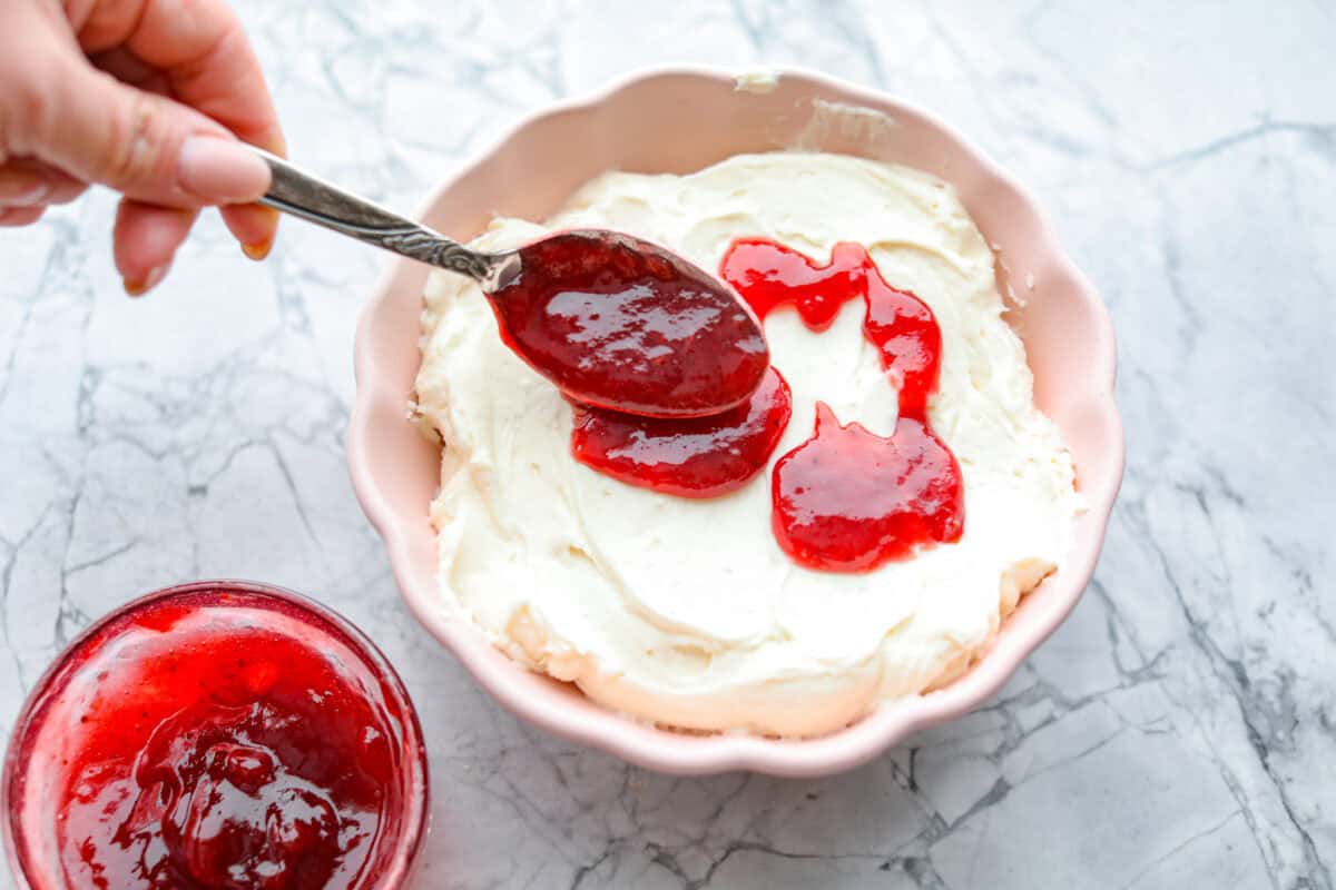 Strawberry cheesecake dip is an irresistible treat that tastes just like the classic but in a fun dip form! Whip it up in just 5 minutes! It's so good with graham crackers, fresh fruit, and cookies for a sweet, crowd-pleasing snack! Overhead shot of someone spreading strawberry jam over the cream cheese mixture.