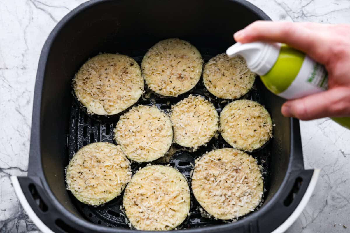 Crispy Air Fryer Eggplant is a fun flavor snack that is a healthier option than typical fried snacks. Tender eggplant slices are coated in crispy breadcrumbs and parmesan cheese, deliciously seasoned, then air-fried to perfection. Overhead shot of someone spraying coated slices in the bottom of the air fryer basket.