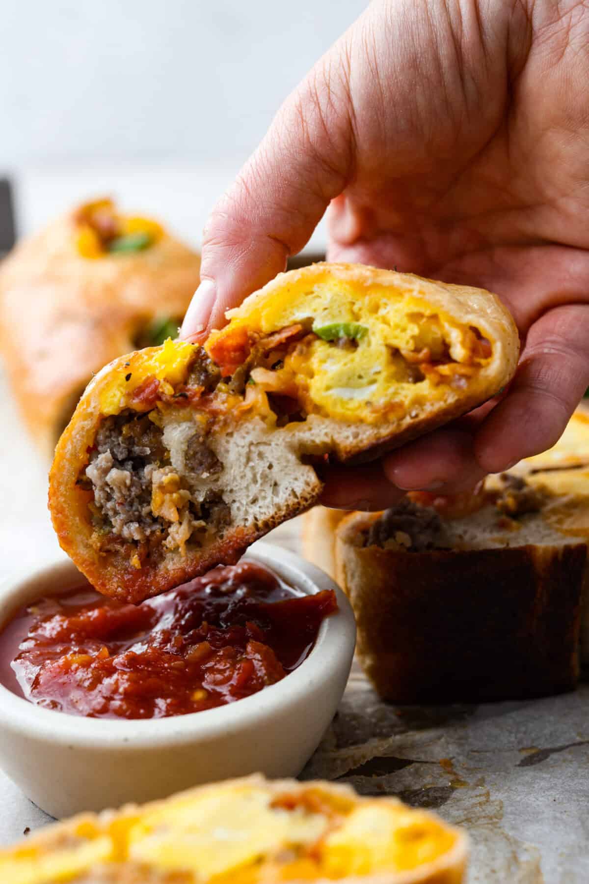 This Breakfast Stromboli is the best morning treat! It's packed with fluffy eggs, savory sausage, crispy bacon, and melty cheese, all wrapped in golden pizza dough. Perfect for holiday brunches or everyday family breakfasts! Side shot of someone dipping a piece of the breakfast Stromboli into salsa.