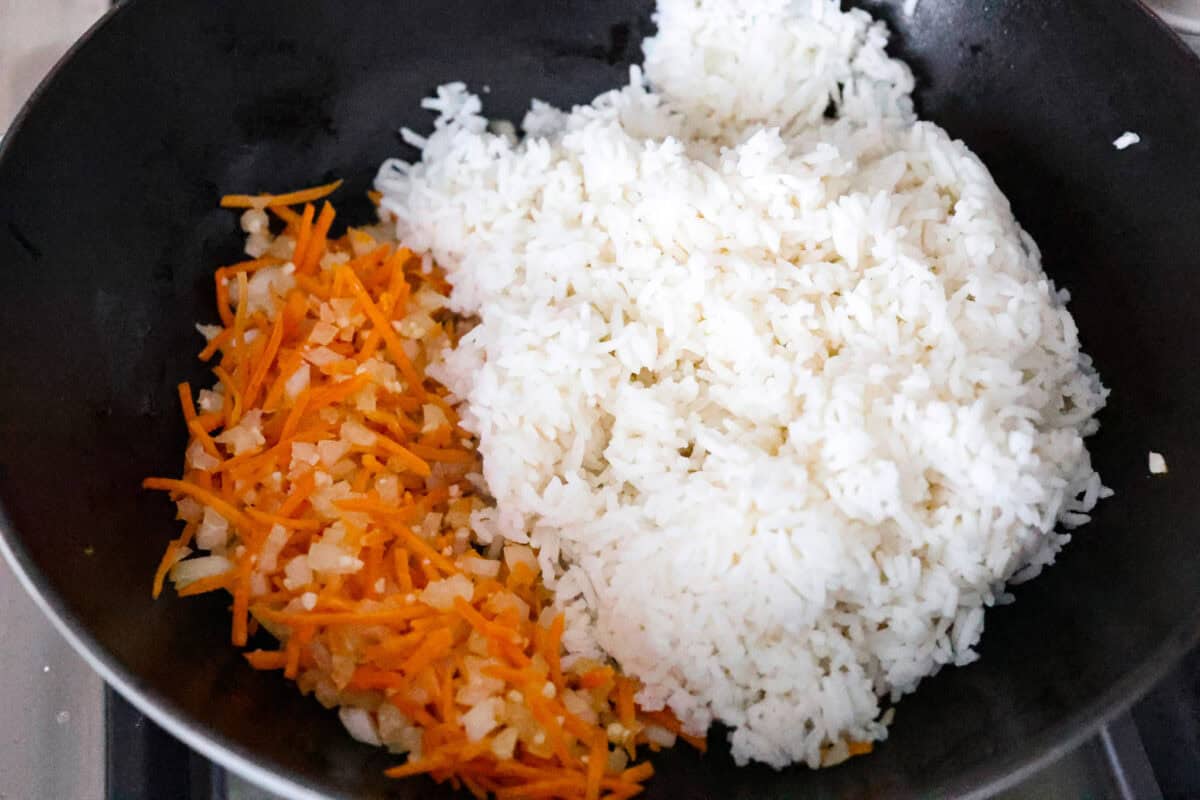 Delicious crab fried rice comes together in just 25 minutes with simple ingredients and tons of flavor! It combines the sweetness of lump crab meat with the savory flavors of classic fried rice for an endlessly satisfying dish that's sure to become a family favorite! Overhead shot of cooked rice being added to the cooked vegetables in the skillet.