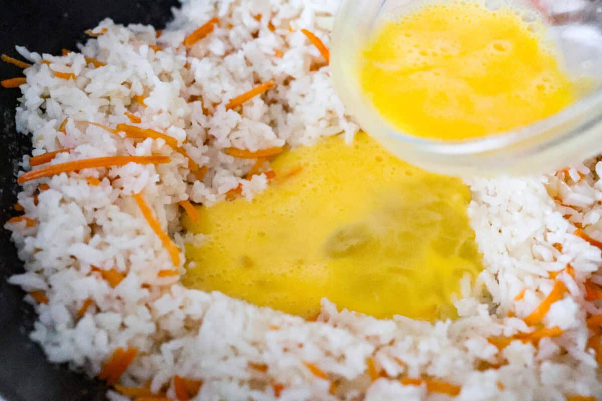 Delicious crab fried rice comes together in just 25 minutes with simple ingredients and tons of flavor! It combines the sweetness of lump crab meat with the savory flavors of classic fried rice for an endlessly satisfying dish that's sure to become a family favorite! Close up shot of someone pouring whisked eggs into a space made in the middle of the rice and veggies in the skillet.