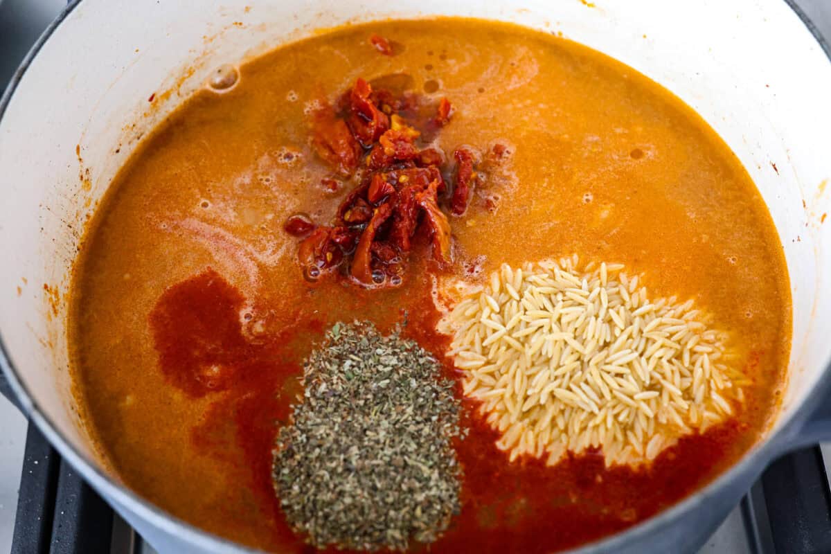Warm up to this delicious Marry Me sausage orzo soup! It's hearty, super flavorful, and packed with seasoned sausage, tender orzo pasta, and plenty of bold, savory seasonings. Overhead shot of chicken broth, seasonings, orzo and sun dried tomatoes added to the sausage and onions.