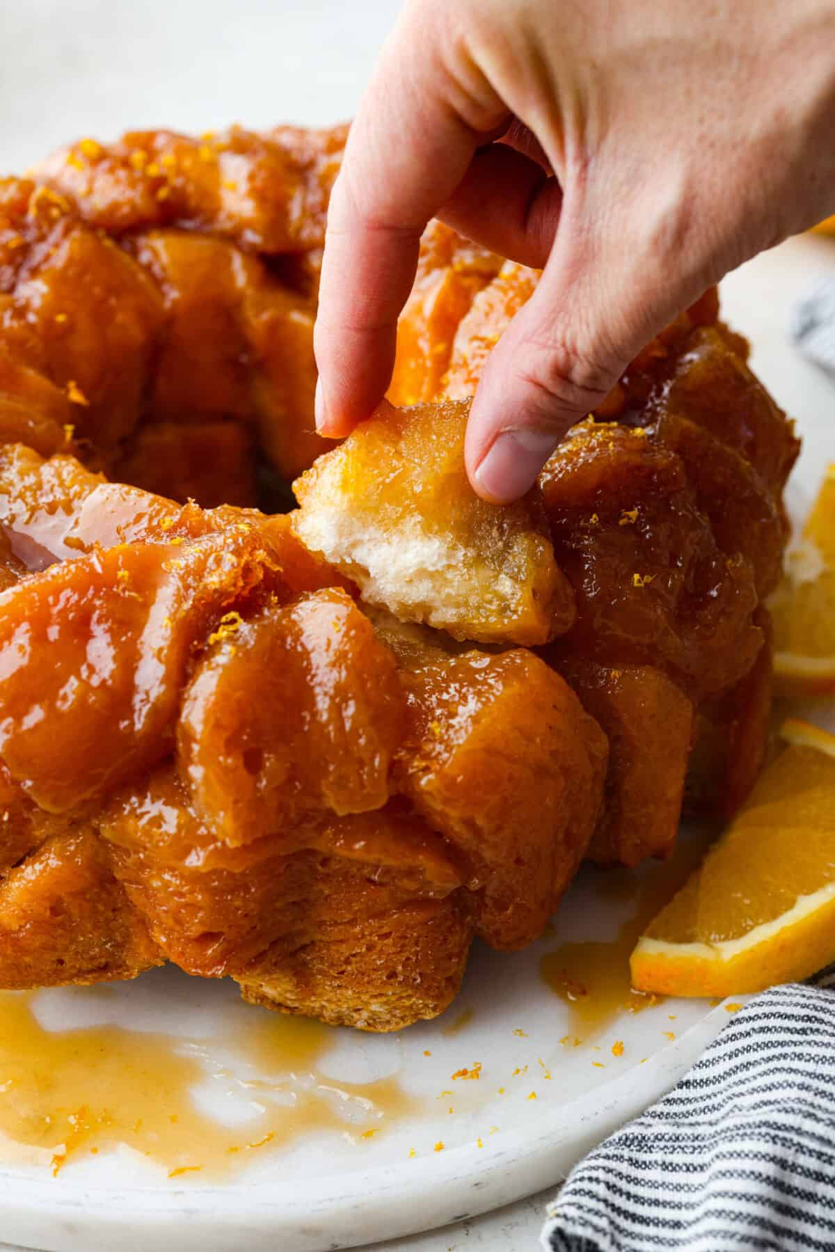 Orange monkey bread is a fun twist on a classic sticky pull-apart bread. It's packed with soft, buttery biscuit pieces, bursts of citrusy orange zest, and a sticky, sweet glaze. Everyone will want to get their hands on this delicious dish! Close up shot of someone pulling a piece of orange monkey bread off the whole bundt.