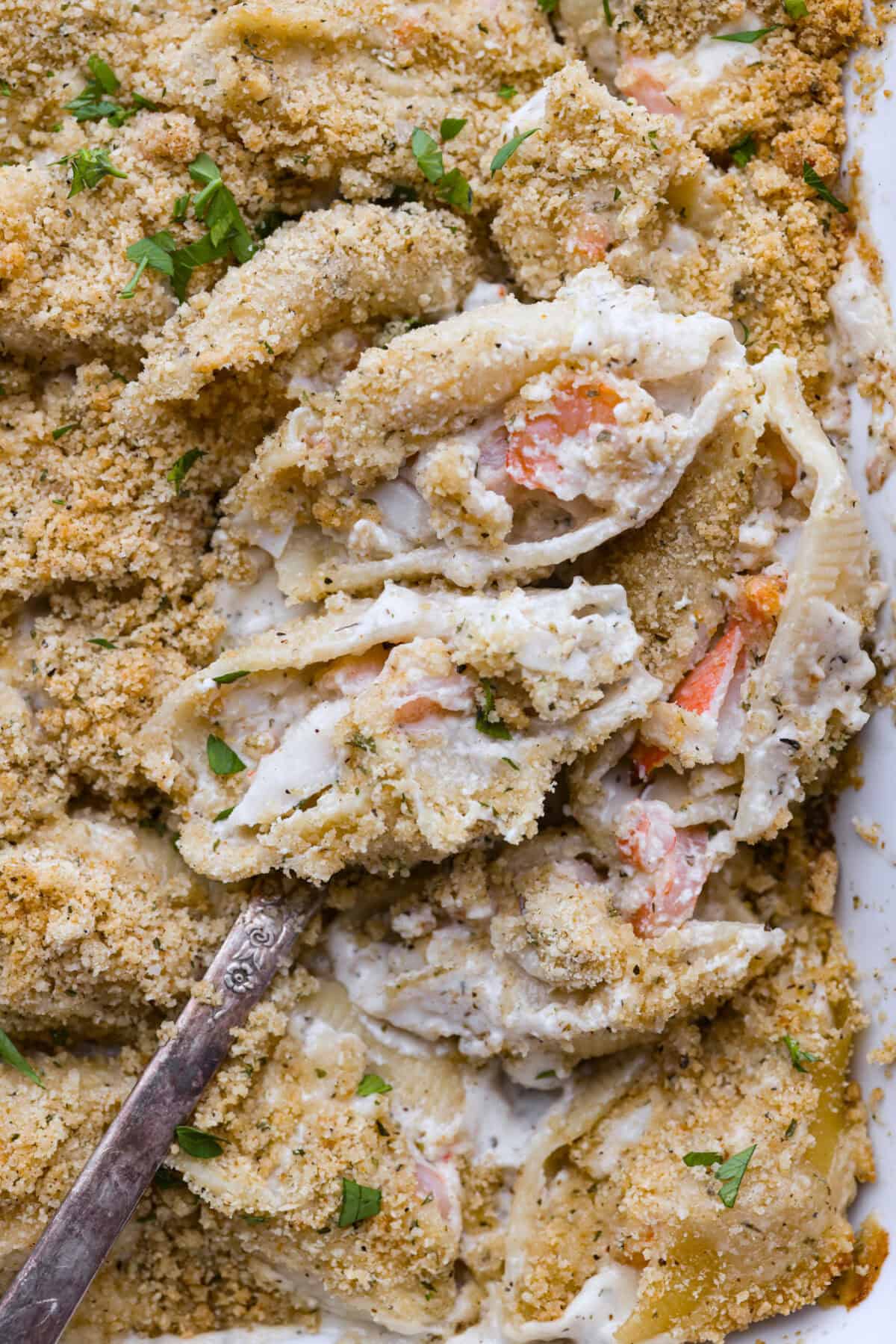 Seafood Stuffed Shells is a comforting meal that's sure to impress! Jumbo pasta shells are generously filled with a creamy, savory seafood filling and coated in a rich Alfredo-based sauce. Finish with a crispy crumb topping for an irresistible dinner! Close up shot of seafood stuffed shells on a serving spoon in the baking dish.