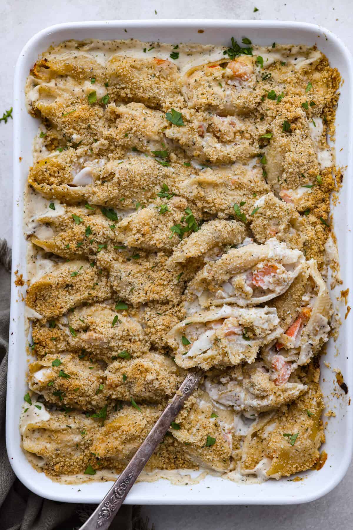 Seafood Stuffed Shells is a comforting meal that's sure to impress! Jumbo pasta shells are generously filled with a creamy, savory seafood filling and coated in a rich Alfredo-based sauce. Finish with a crispy crumb topping for an irresistible dinner! Overhead shot of a baking dish with seafood stuffed shells and a serving spoon.