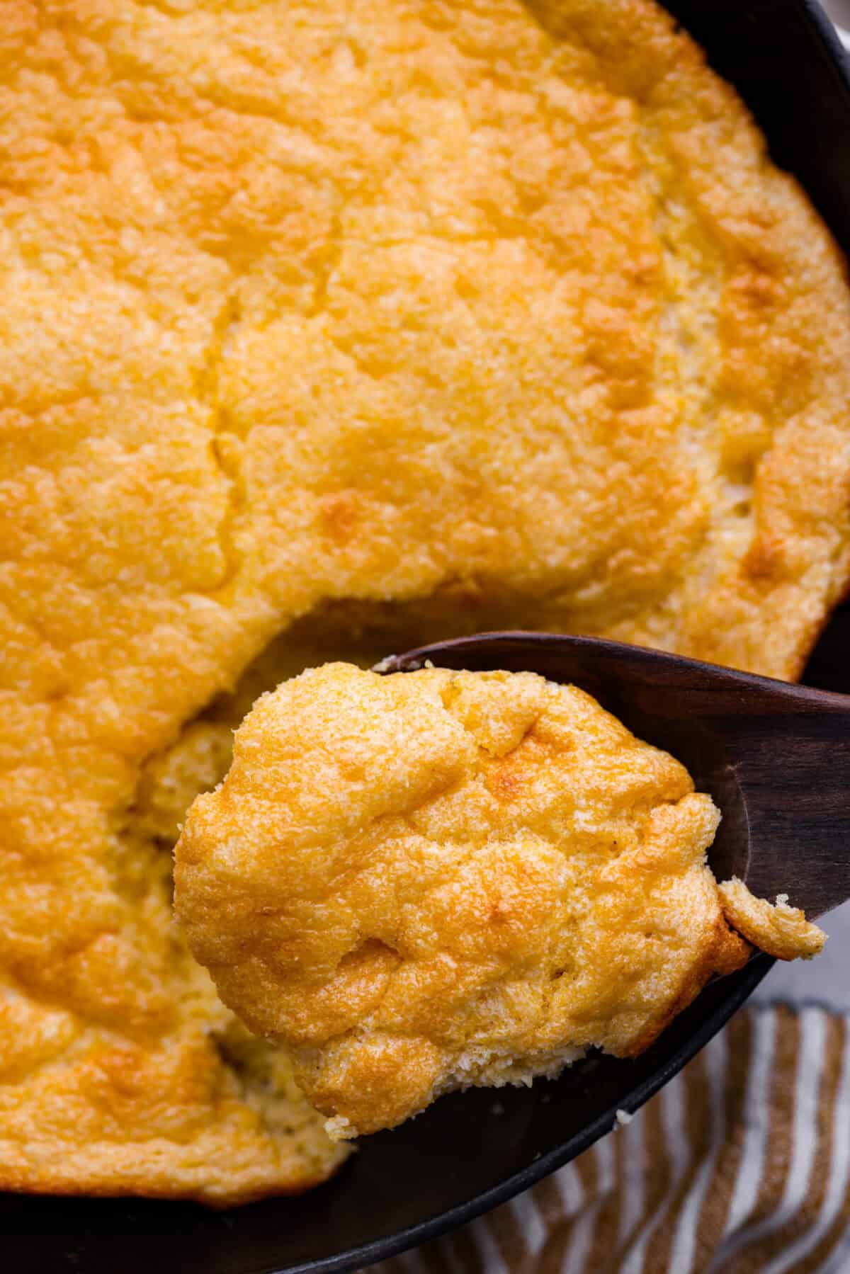Spoon bread is a beloved classic Southern side dish. It’s similar to cornbread but is less sweet and has a softer, almost soufflé-like texture. This unique bread is best enjoyed warm with a dollop of butter, a drizzle of honey or served alongside a hearty bowl of chili. Close up shot of a scoop of spoon bread in a wooden spoon over the skillet.