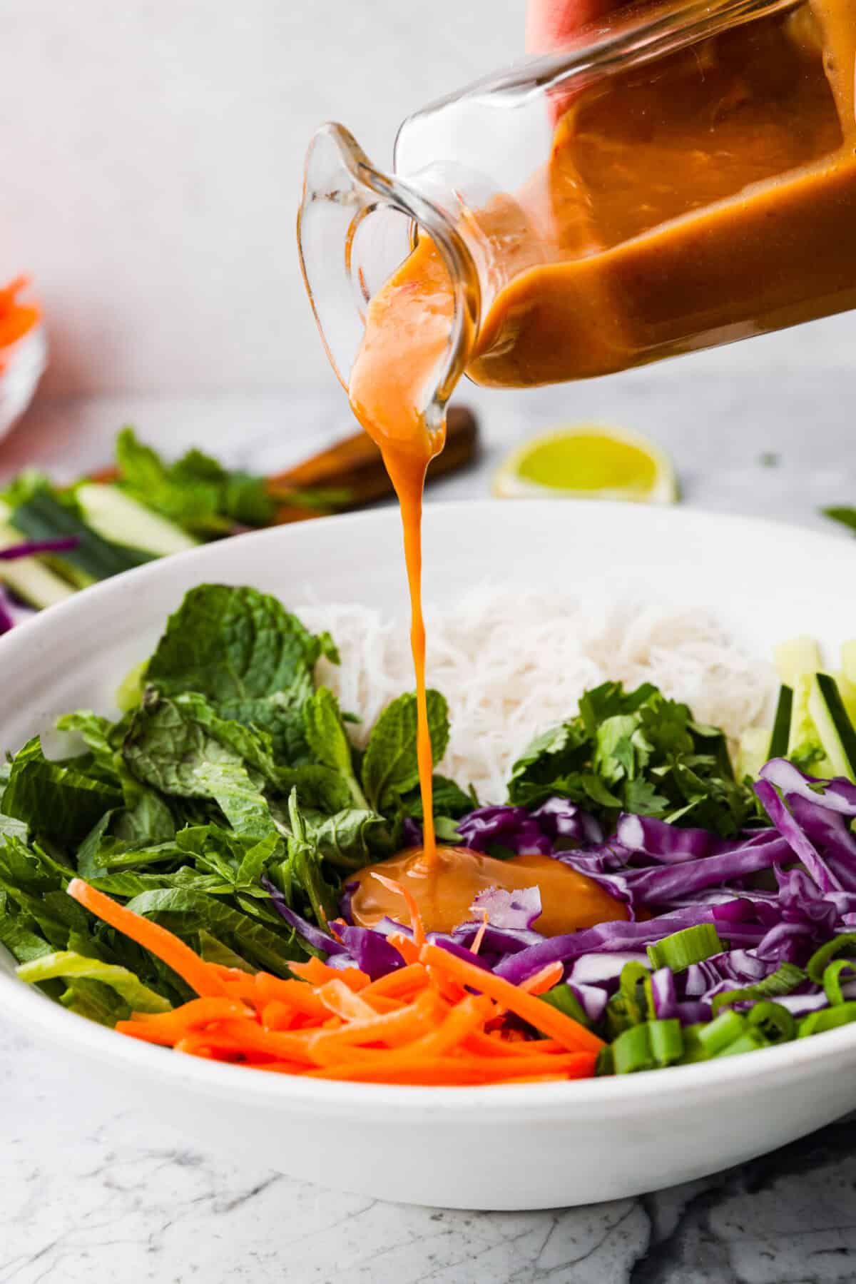 This flavorful, fresh Spring Roll Bowl is not only insanely delicious but absolutely beautiful. It's filled with tender rice vermicelli, vibrant vegetables, and fresh herbs, all topped with my Thai peanut dressing. Side shot of someone pouring Thai peanut dressing into the spring roll bowl.