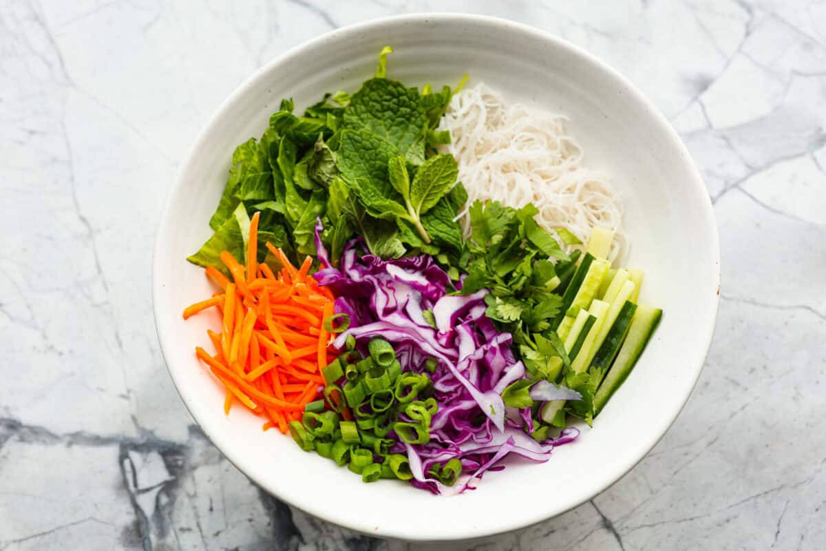 This flavorful, fresh Spring Roll Bowl is not only insanely delicious but absolutely beautiful. It's filled with tender rice vermicelli, vibrant vegetables, and fresh herbs, all topped with my Thai peanut dressing. Overhead shot of a bowl with carrots, cabbage, cucumber, onion, cilantro lettuce, and mint on top of the noodles.