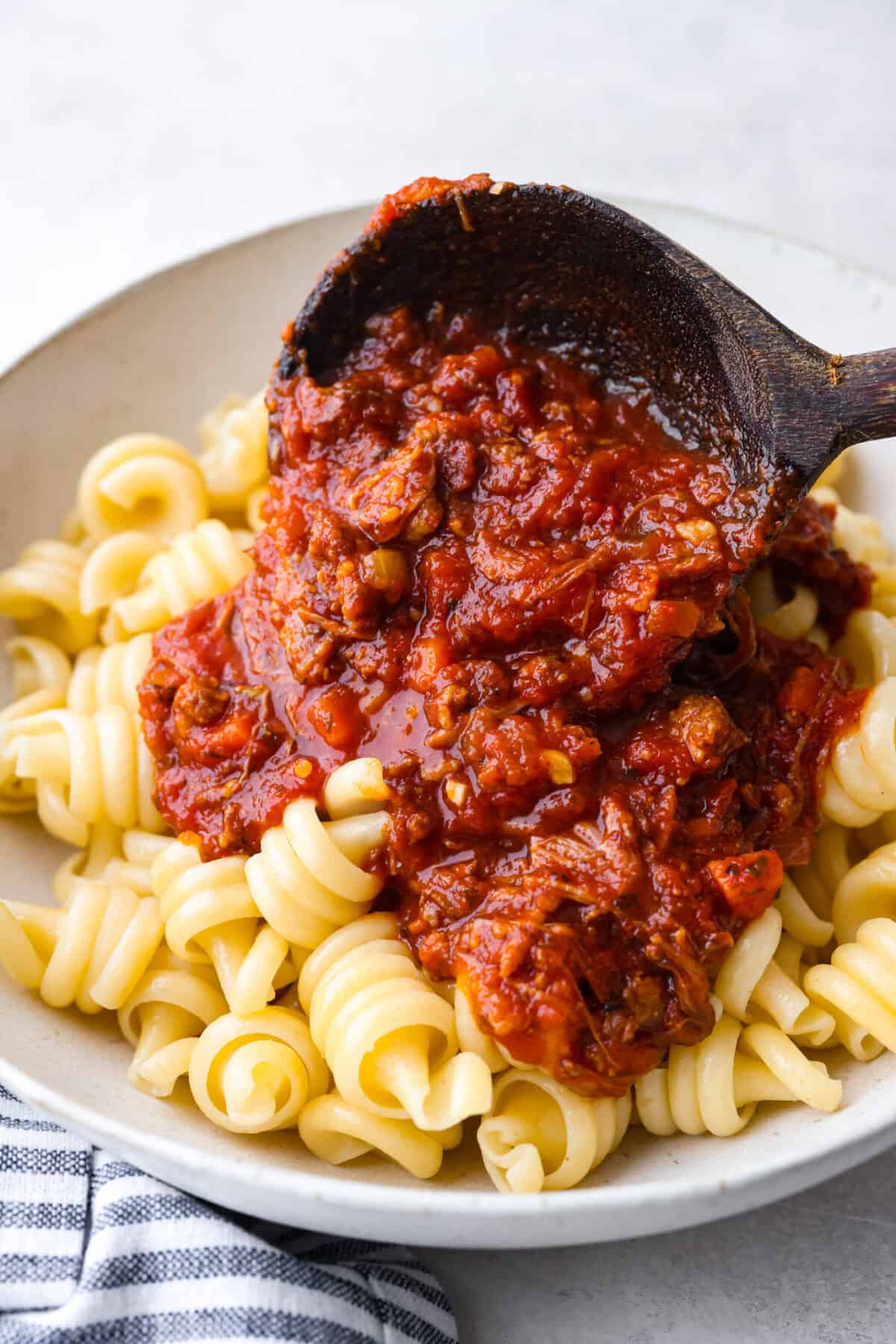 Slow cooker Sunday sauce, also known as "sugo," is a rich, tomato-based ragu that’s a favorite for family dinners. Traditionally, it's simmered low and slow from morning until dinnertime, making it a perfect recipe for the Crockpot. Serve it over pasta with a sprinkle of pecorino romano cheese for an unforgettable meal! Side shot of someone pouring a scoop of slow cooker Sunday sauce over a plate of cooked pasta.