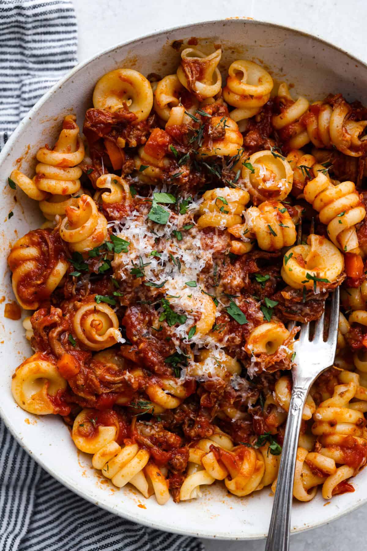 Slow cooker Sunday sauce, also known as "sugo," is a rich, tomato-based ragu that’s a favorite for family dinners. Traditionally, it's simmered low and slow from morning until dinnertime, making it a perfect recipe for the Crockpot. Serve it over pasta with a sprinkle of pecorino romano cheese for an unforgettable meal! Overhead shot of plated slow cooker Sunday sauce with pasta and garnished with parmesan cheese.