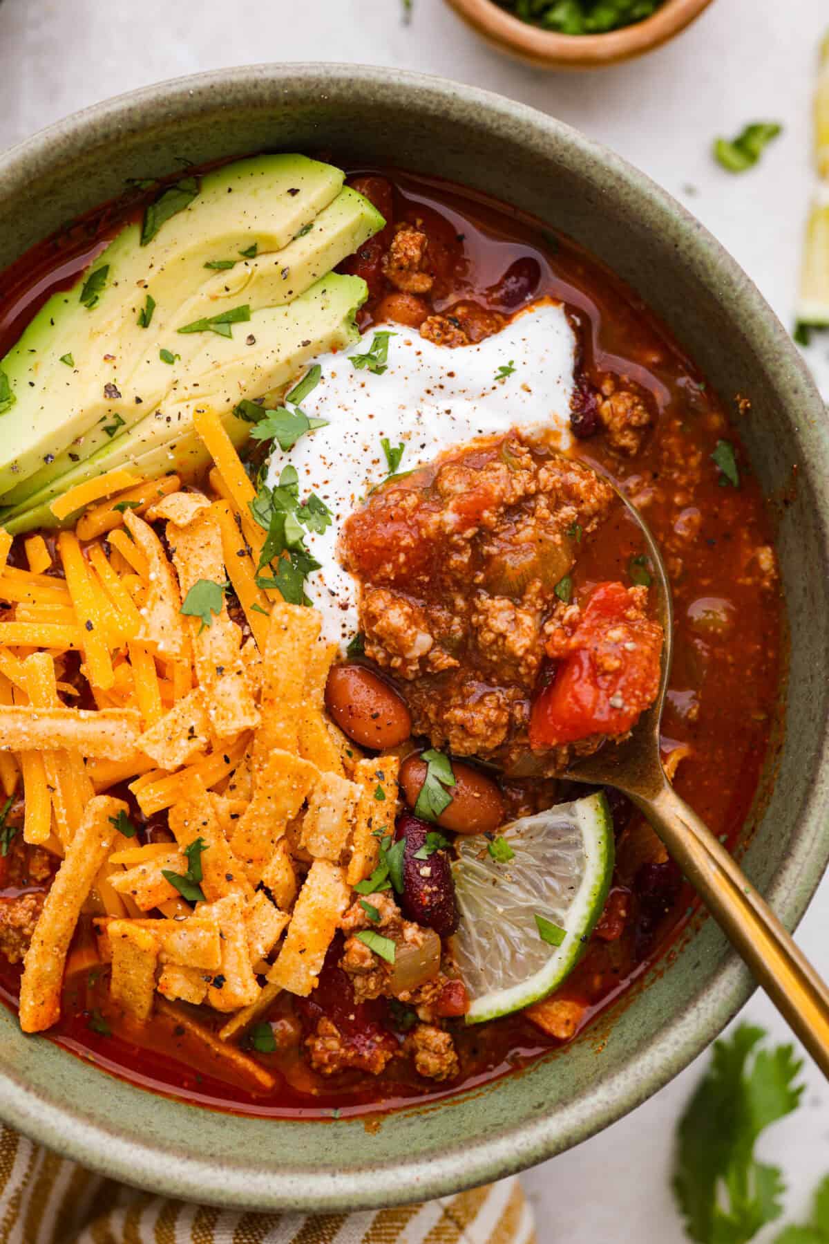 Turkey chili is such a comforting, satisfying meal! Packed with ground turkey, tender vegetables, hearty beans and a blend of bold spices, itโs filled with the most delicious flavor. Great for making ahead and feeding a crowd! Close up shot of a bowl of turkey chili with avocado, lime, cheese sour cream and tortilla strips for garnish.