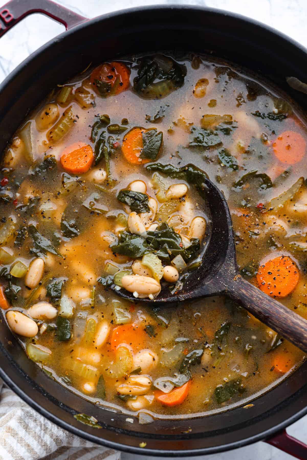 Tuscan white bean soup is like a warm hug in a bowl. Hearty beans and tender vegetables come together to give you a boost of nutrients. This soup is light and fresh tasting, with bright, zesty flavors! Overhead shot of Tuscan white bean soup in a pot with a wooden spoon.