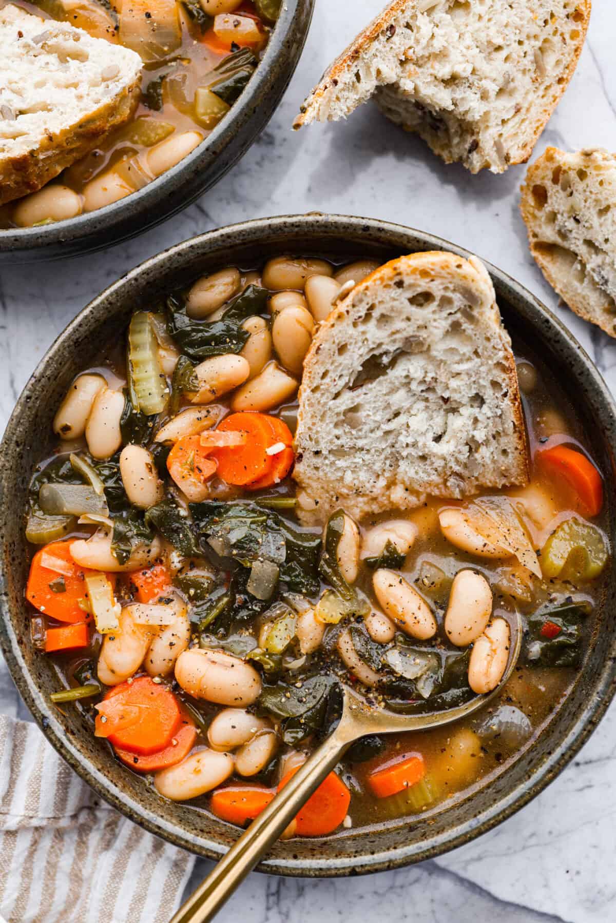 Tuscan white bean soup is like a warm hug in a bowl. Hearty beans and tender vegetables come together to give you a boost of nutrients. This soup is light and fresh tasting, with bright, zesty flavors! Overhead shot of a bowl of Tuscan white bean soup with a half a slice of bread in it.