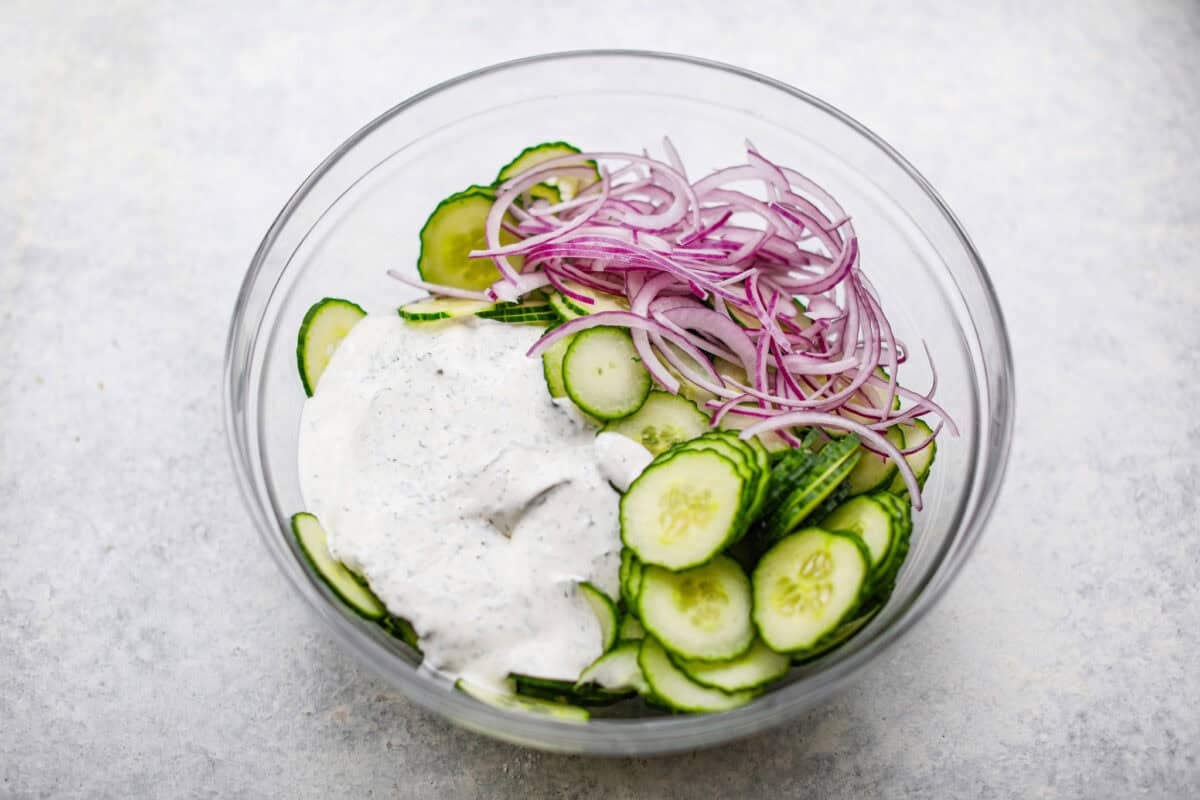 This refreshing German cucumber salad, or Gurkensalat, is easy to throw together and is so flavorful. Thinly sliced cucumbers and fresh herbs are infused in a creamy, tangy sauce. It makes the perfect healthy snack or side dish. Overhead shot of sliced cucumbers, red onions and sauce in a bowl.