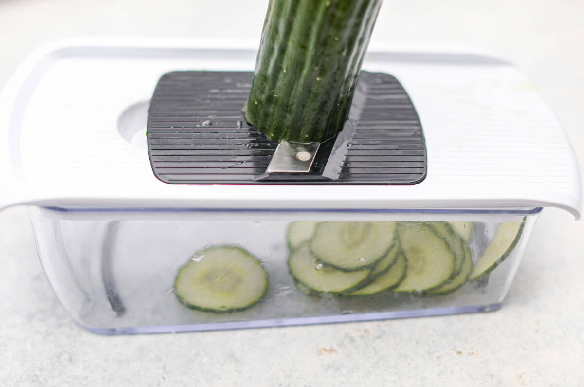 This refreshing German cucumber salad, or Gurkensalat, is easy to throw together and is so flavorful. Thinly sliced cucumbers and fresh herbs are infused in a creamy, tangy sauce. It makes the perfect healthy snack or side dish. Angles hot of someone slicing a cucumber using a Mandoline.
