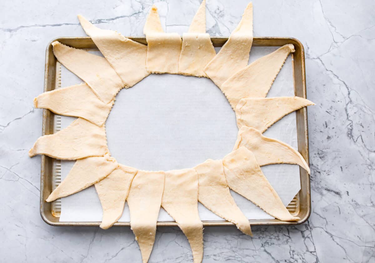 This Raspberry Cream Cheese Crescent Ring is a simple pastry with a flaky, golden crust, a sweet and tangy cream cheese filling, and a burst of raspberry flavor. Perfect for breakfast, brunch, or dessert, it's easy to make yet impressively delicious! Overhead shot of a baking sheet with parchment paper and crescent rolls spread out in a sunburst shape.