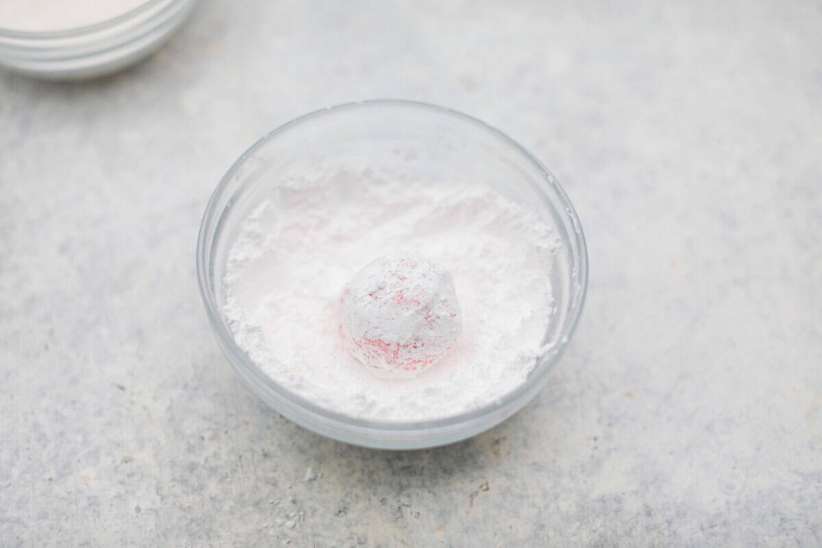 Strawberry Crinkle Cookies are the perfect blend of soft and chewy with a sweet, powdered sugar coating. Their vibrant color and classic strawberry cake flavor make them perfect for any occasion. Angle shot of a ball of dough rolled in a bowl of powdered sugar.