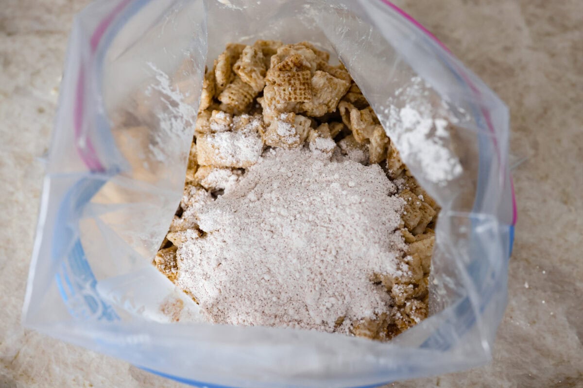 Churro Chex Mix is a fun variation of the popular muddy buddy mix. Sweet cinnamon and sugar mixed with silky white chocolate coat the crunchy cereal to create an addicting treat. It's the perfect snack to whip up for game day, road trips, or weeknights when that sweet tooth hits! Overhead shot of the chocolate covered cereal in a large ziplock with the cinnamon and sugar mixture poured over the top.