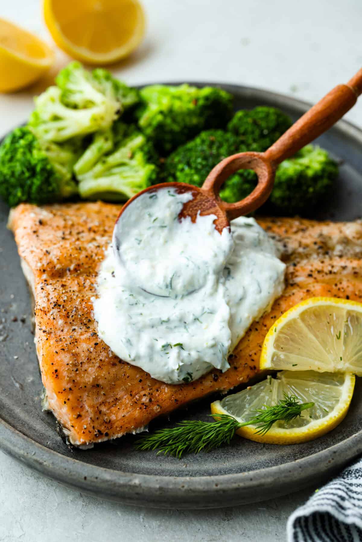 This creamy lemon dill sauce is fresh, tangy, and packed with flavor! Itโs the perfect complement to salmon, roasted veggies, or even as a dip for crispy potatoes. Made with simple ingredients like fresh dill, lemon, and a touch of garlic, it comes together in minutes! Angle shot of someone spreading fresh lemon dill sauce over a fillet of plated fish and broccoli, garnished with lemon wedges and fresh dill.