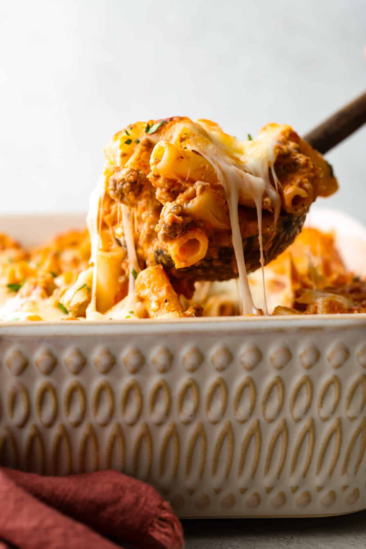 Million Dollar Baked Ziti is the ultimate comfort food that tastes like a million bucks! It's packed with layers of tender noodles, ground meat, spaghetti sauce, and plenty of cheesy goodness. There is something for the whole family in this hearty, crowd-pleasing dish! SIde shot of someone scooping a portion of million dollar baked ziti out of the casserole dish.