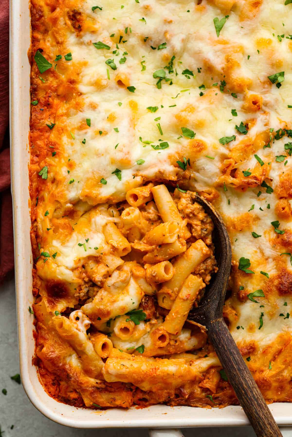 Million Dollar Baked Ziti is the ultimate comfort food that tastes like a million bucks! It's packed with layers of tender noodles, ground meat, spaghetti sauce, and plenty of cheesy goodness. There is something for the whole family in this hearty, crowd-pleasing dish! Overhead shot of million dollar baked ziti with a wooden serving spoon getting a scoop out.