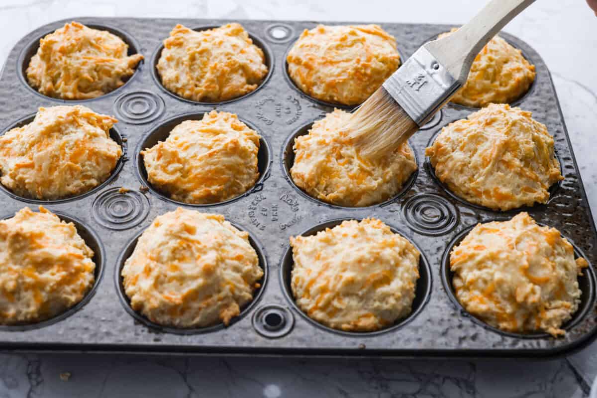 These cheesy beer bread muffins are light, fluffy, and packed with savory flavor! The key is to not overmix the batter. Mixing until just combined will give you the perfect airy, melt-in-your-mouth texture that will keep you coming back for more. Angle shot of someone brushing butter over the top of the baked muffins.