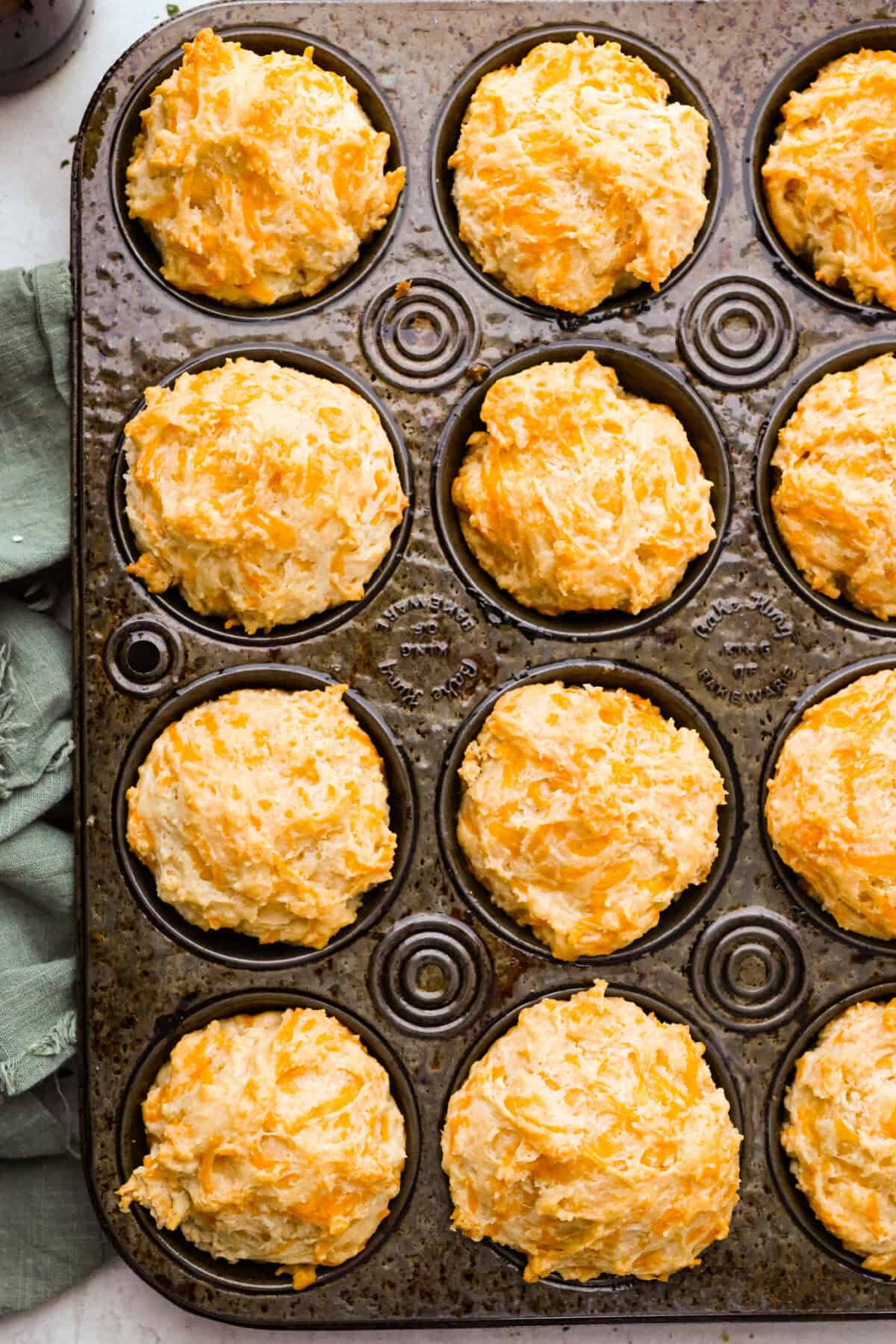 These cheesy beer bread muffins are light, fluffy, and packed with savory flavor! The key is to not overmix the batter. Mixing until just combined will give you the perfect airy, melt-in-your-mouth texture that will keep you coming back for more. Overhead shot of the baked cheesy beer bread muffins still in the baking tin.