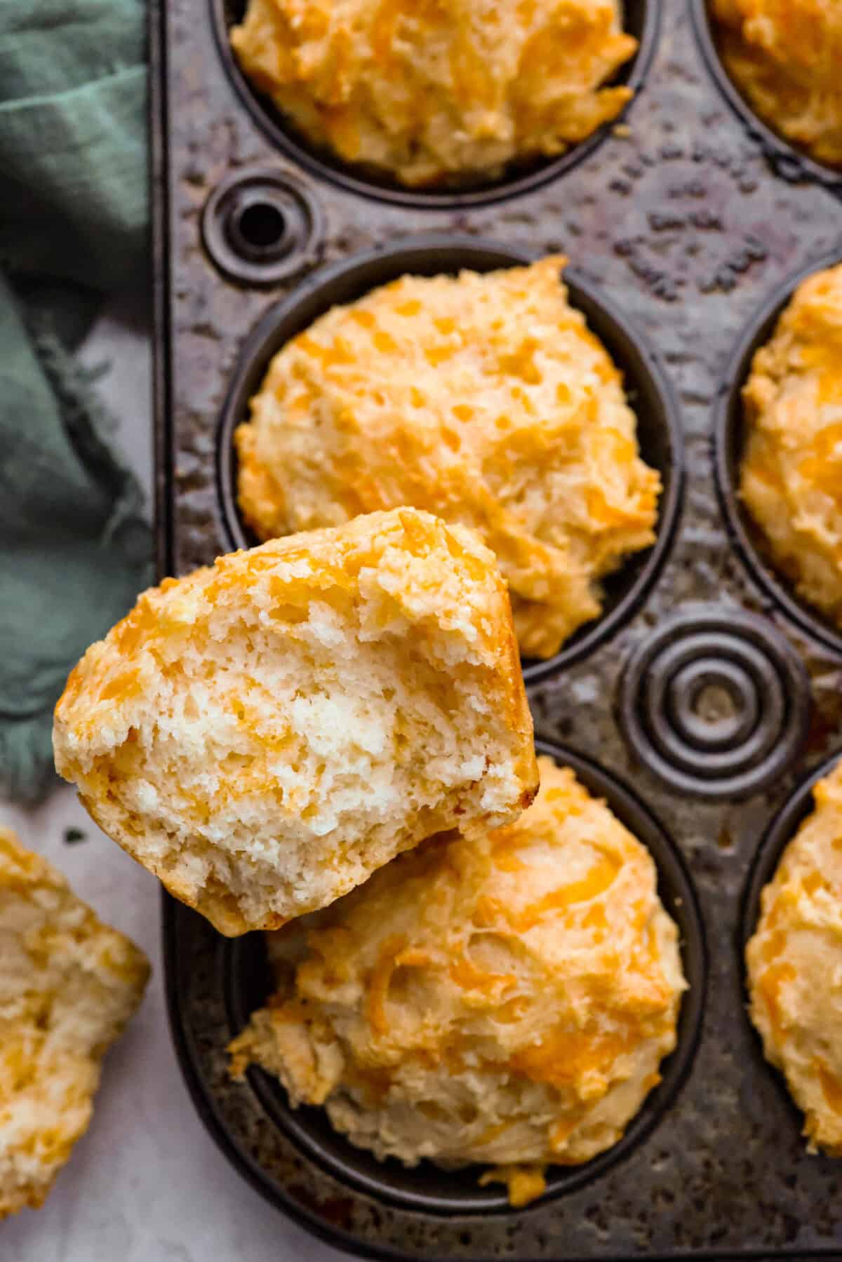 These cheesy beer bread muffins are light, fluffy, and packed with savory flavor! The key is to not overmix the batter. Mixing until just combined will give you the perfect airy, melt-in-your-mouth texture that will keep you coming back for more. Close up shot of a cheesy beer bread muffin torn in half resting on top of the muffins in the muffin tin.
