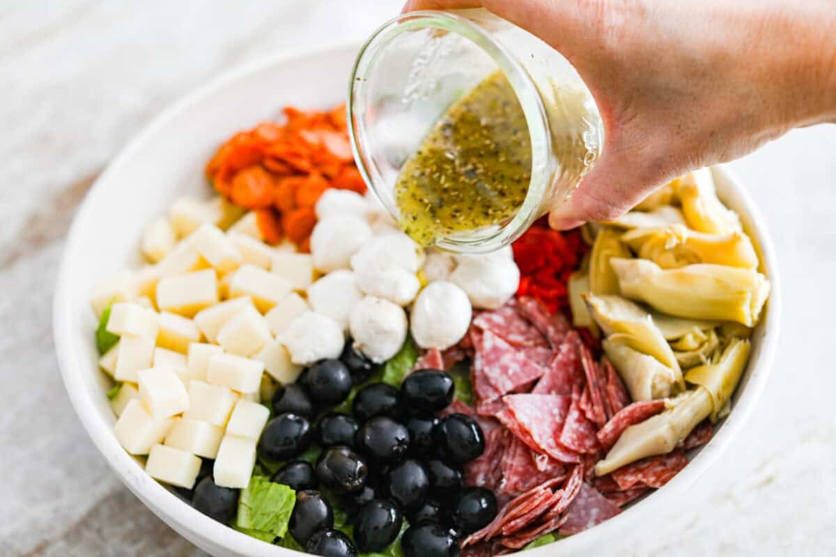 Okay, this antipasto salad? Itโs basically an Italian snack board in a bowl, and Iโmย obsessed! What makes this one a little different (and so good) is the easy lemony dressing. Someone pouring the dressing over the salad ingredients.