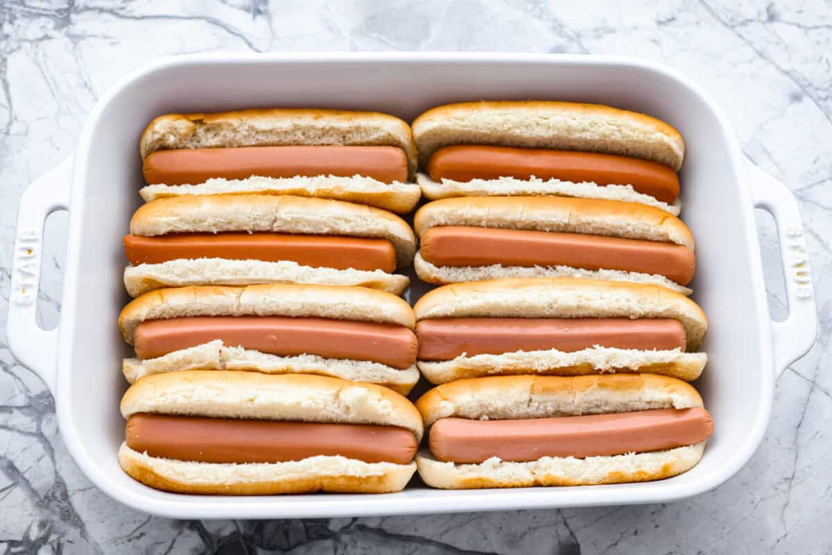 These Baked Chili Cheese Dogs are dangerously good. We’re talking soft buns, hearty chili, and ooey gooey cheese. They’re quick, messy (in the best way), and totally delicious! Hot dogs inside of hot dog buns placed in a baking dish.
