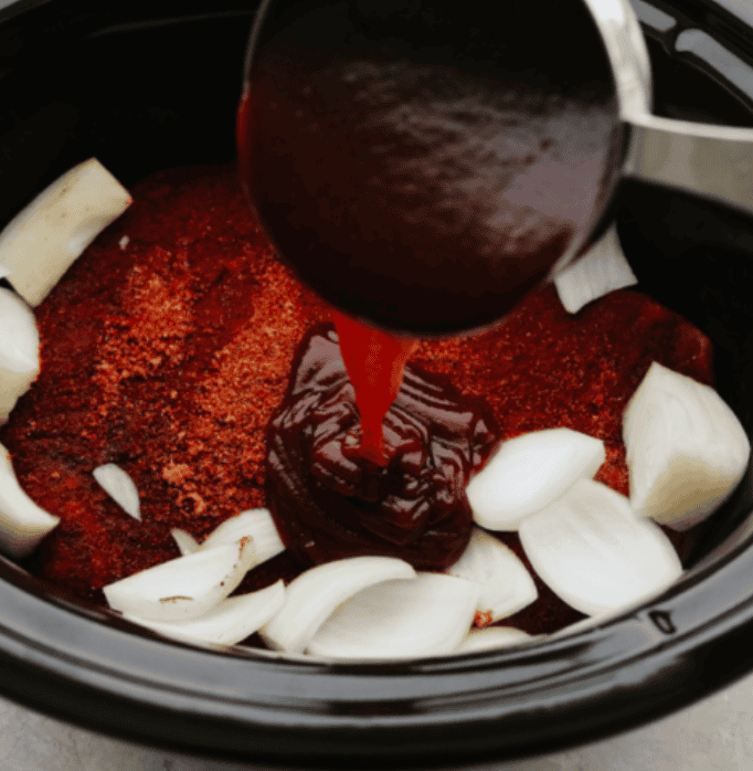 This Slow Cooker Beef Brisket is melt-in-your-mouth tender, packed with flavor, and practically cooks itself. Perfect for easy family dinners or feeding a hungry crowd! Someone pouring bbq sauce over the seasoned meat and onions in the crockpot.