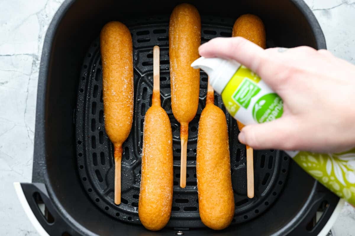 Air fryer corn dogs bring fair-style crunch without the extra oil! Crispy outside, juicy inside, and ready in minutes—perfect for a quick snack or easy meal! Someone spraying corn dogs with oil spray.